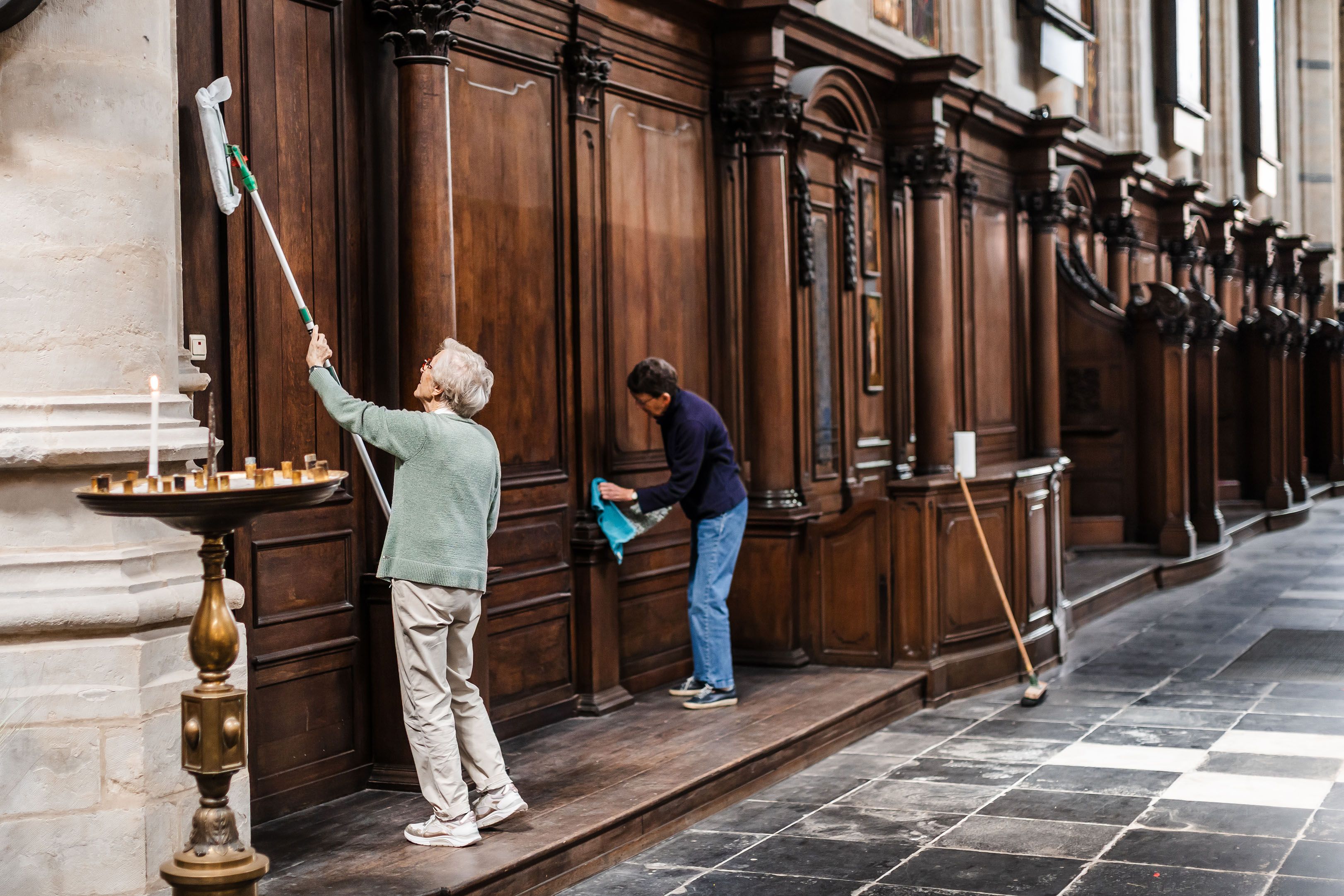 Persoon maakt een houten kerkmeubel schoon of restaureert het interieur van een kerk. Op de achtergrond zijn donkere houten panelen en kerkbanken zichtbaar. De ruimte heeft een stenen vloer en hoge plafonds.