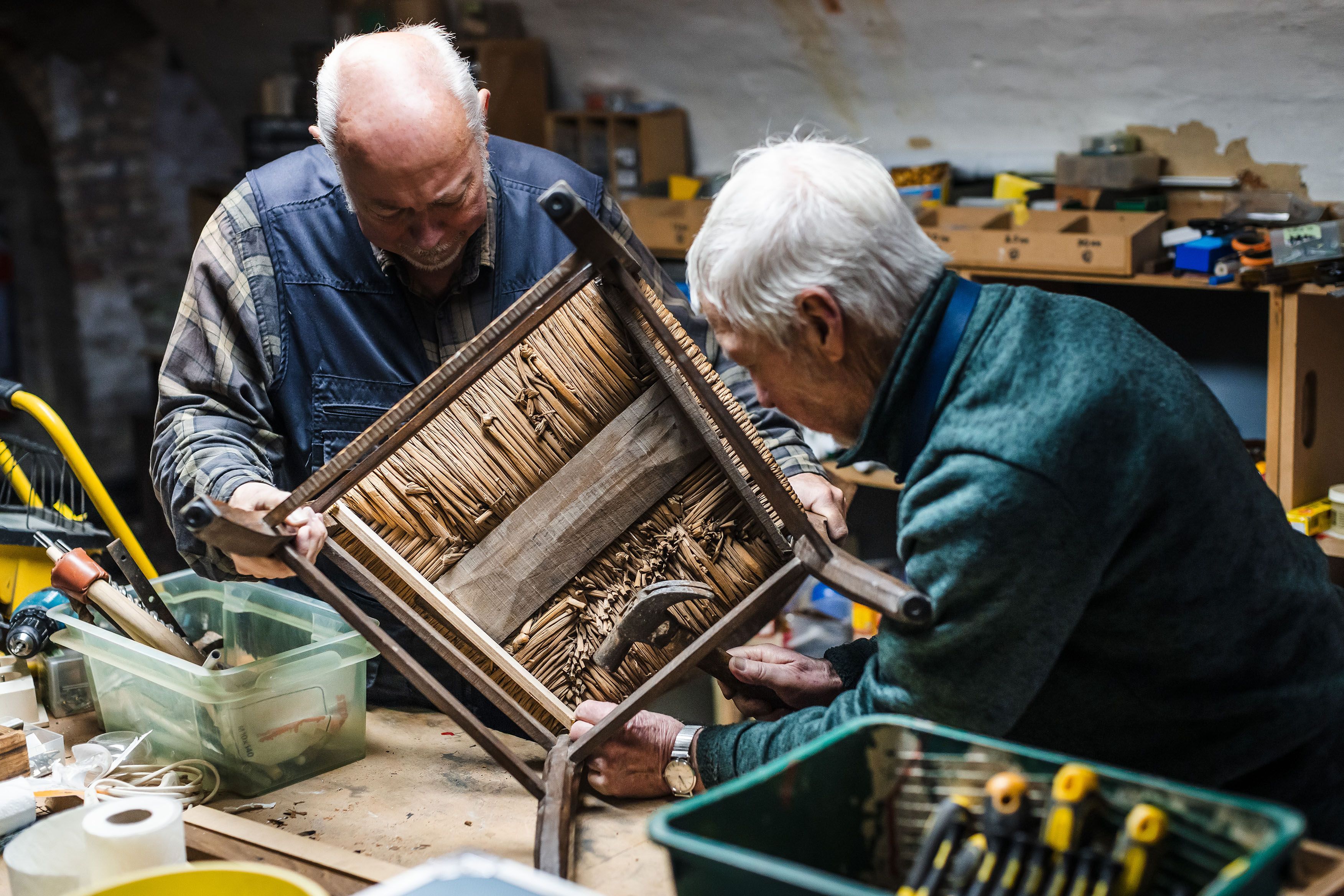 Twee personen restaureren een kunstwerk of schilderijlijst in een werkplaats. Het object ligt op een werktafel, omringd door gereedschap, materialen en restauratiebenodigdheden.