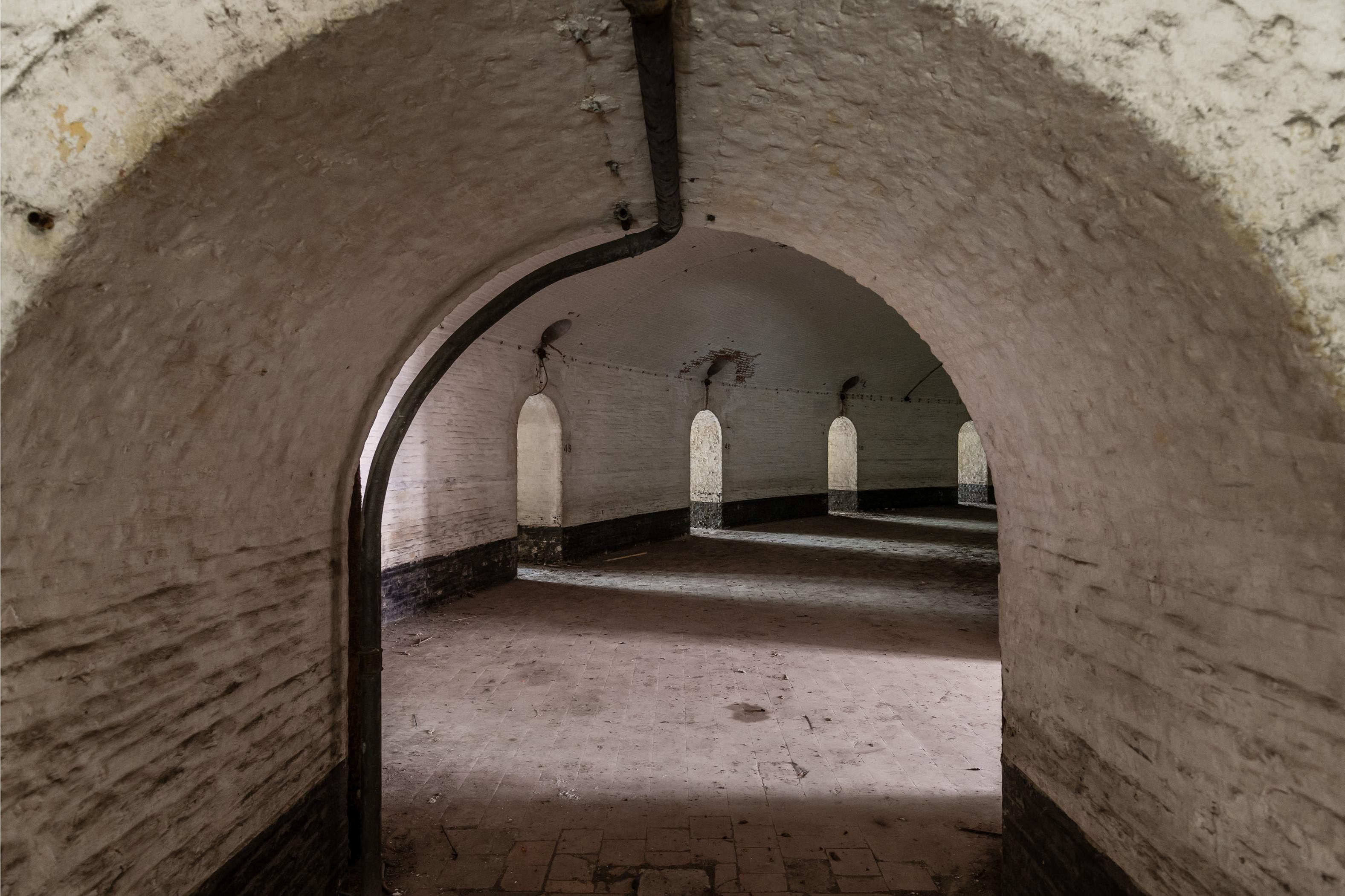 Witgekalkte gebogen gang in fort 7. De ruimte wordt verlicht door boogvormige muuropeningen.