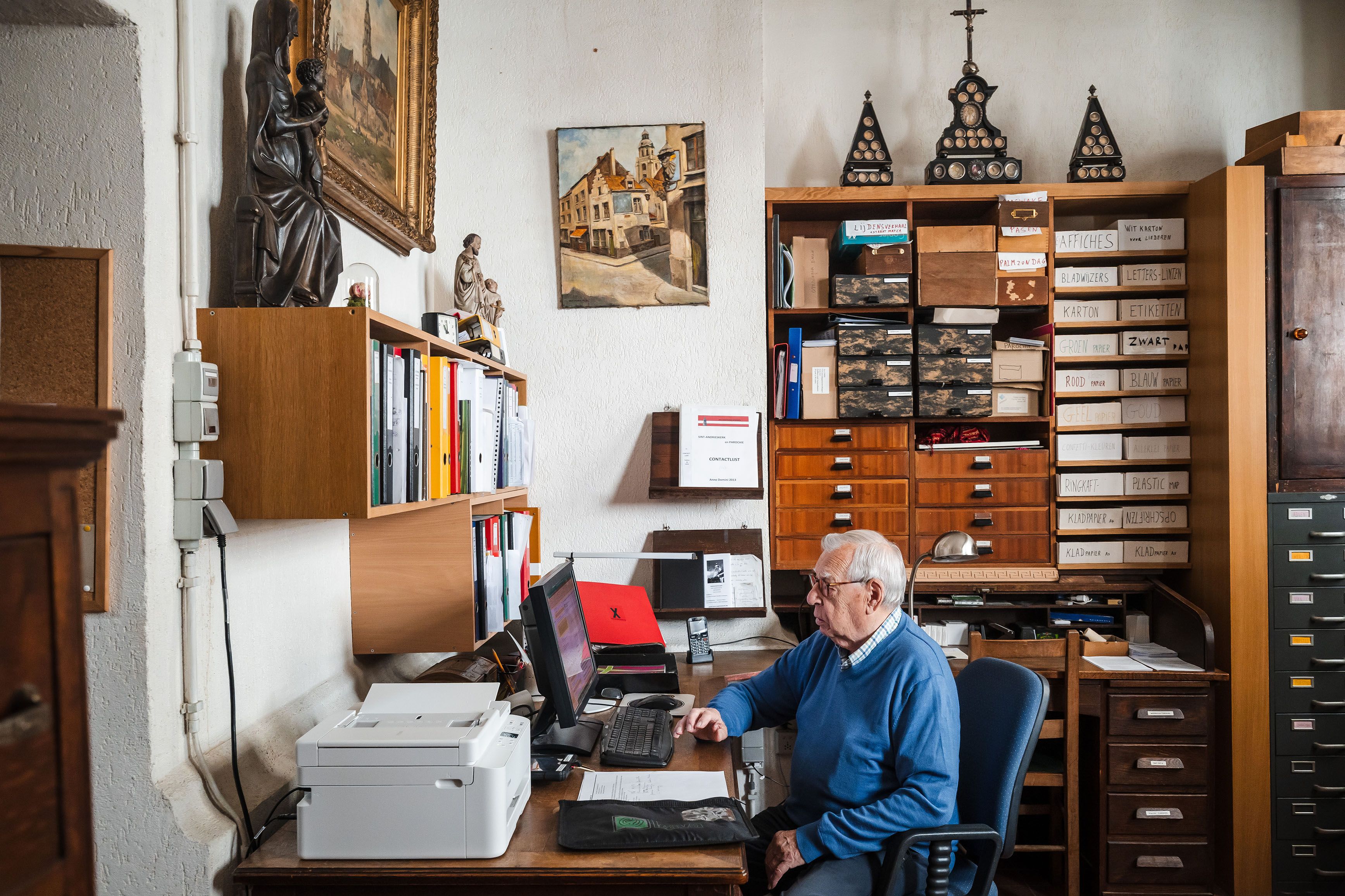 Persoon werkt aan een bureau in een archief- of kantoorruimte. Rondom staan kasten met documenten, boeken en mappen. Op de achtergrond zijn historische voorwerpen en archiefmateriaal zichtbaar.