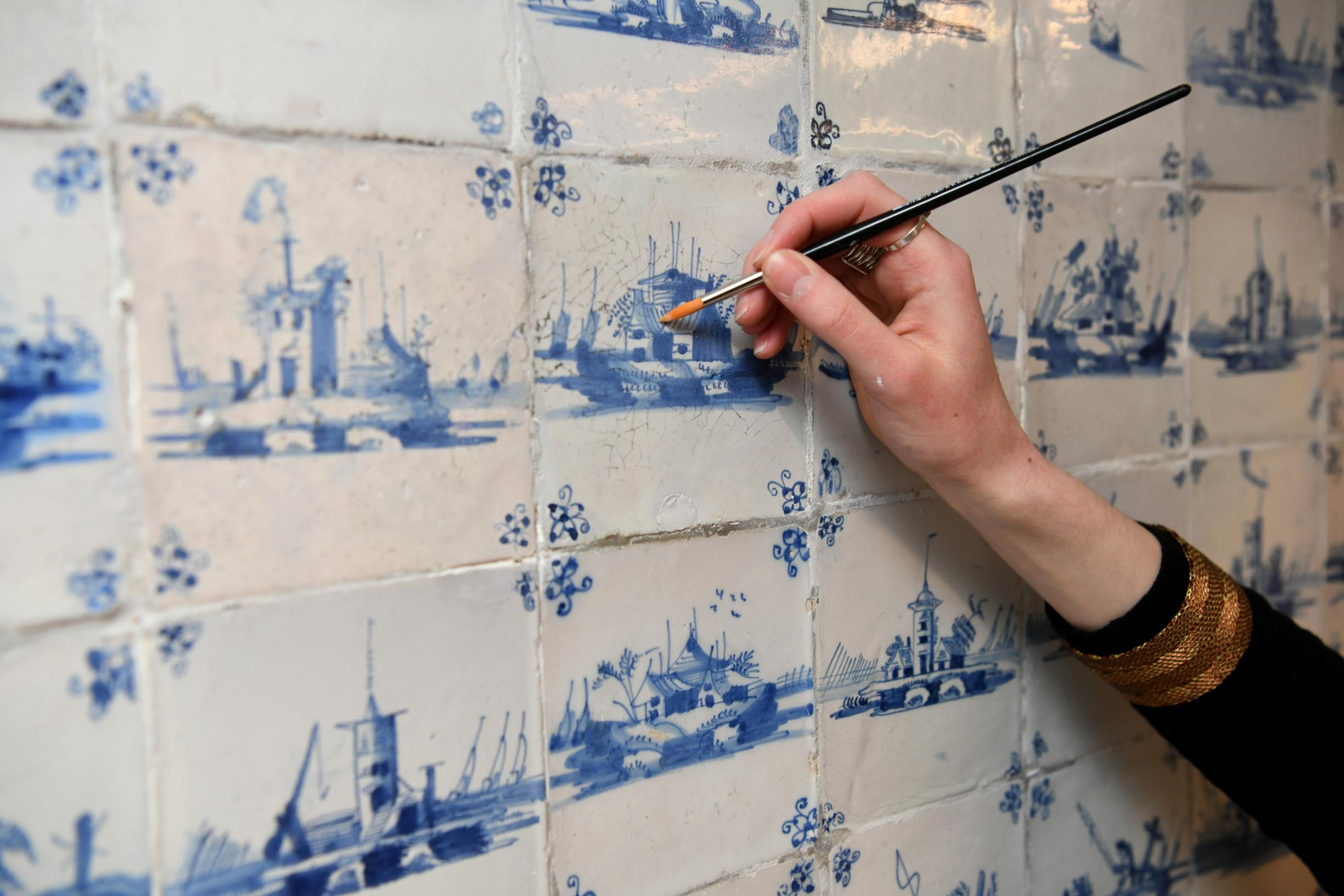 De foto toont een vrouwenhand met een fijn penseel terwijl ze retouches aanbrengt aan een blauw-witte tegel in de kelderkeuken.