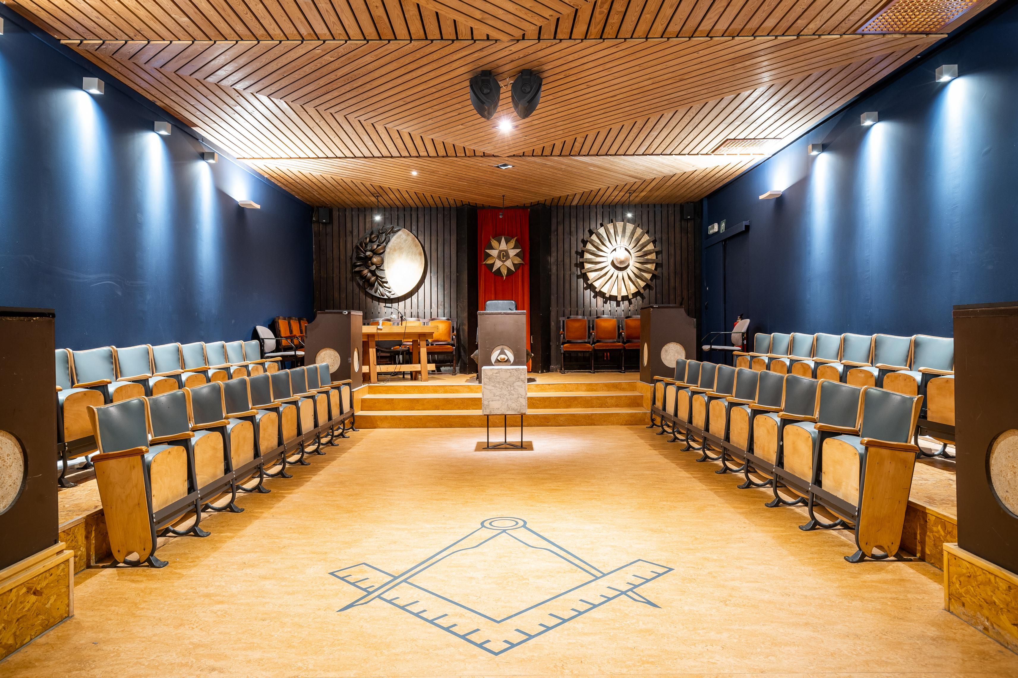 Beeld van vrijmetselarijtempel. Op de grond een passer en winkelhaak en langs beide kanten een rij met stoelen. Vooraan een sprekersgestoelte.