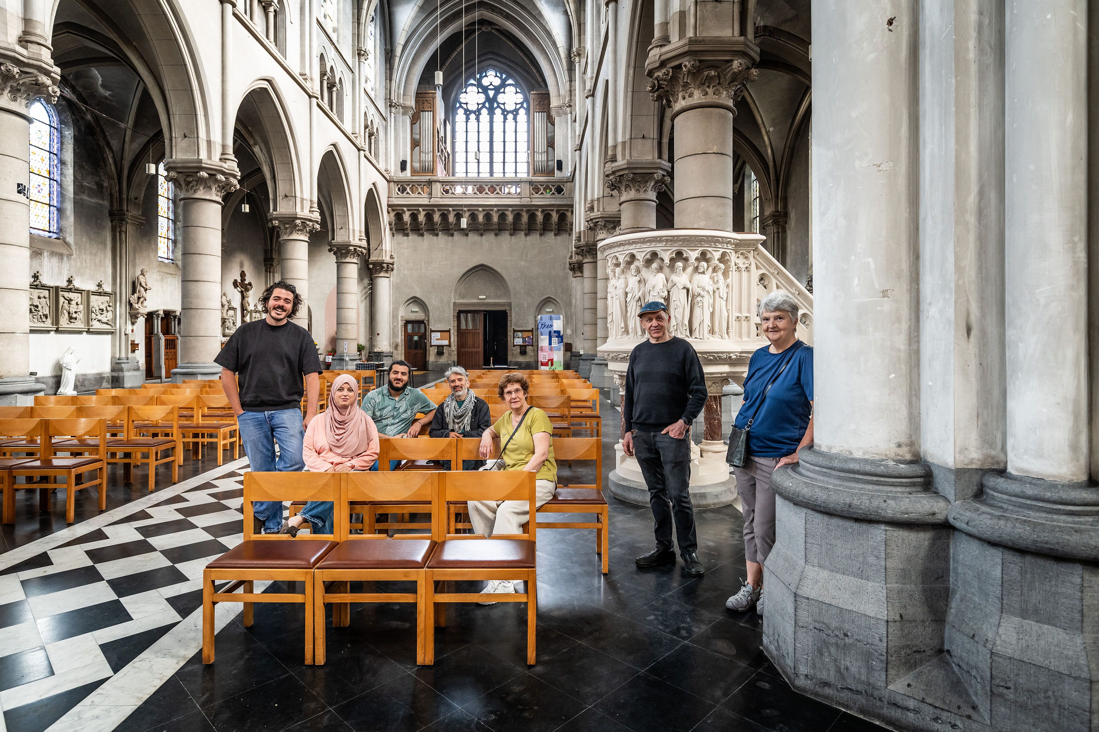 7 vrijwilligers poseren in de Heilige Familiekerk, 2 mannen en 1 vrouw staan recht, 2 mannen en 2 vrouwen zitten op een houten kerkstoel
