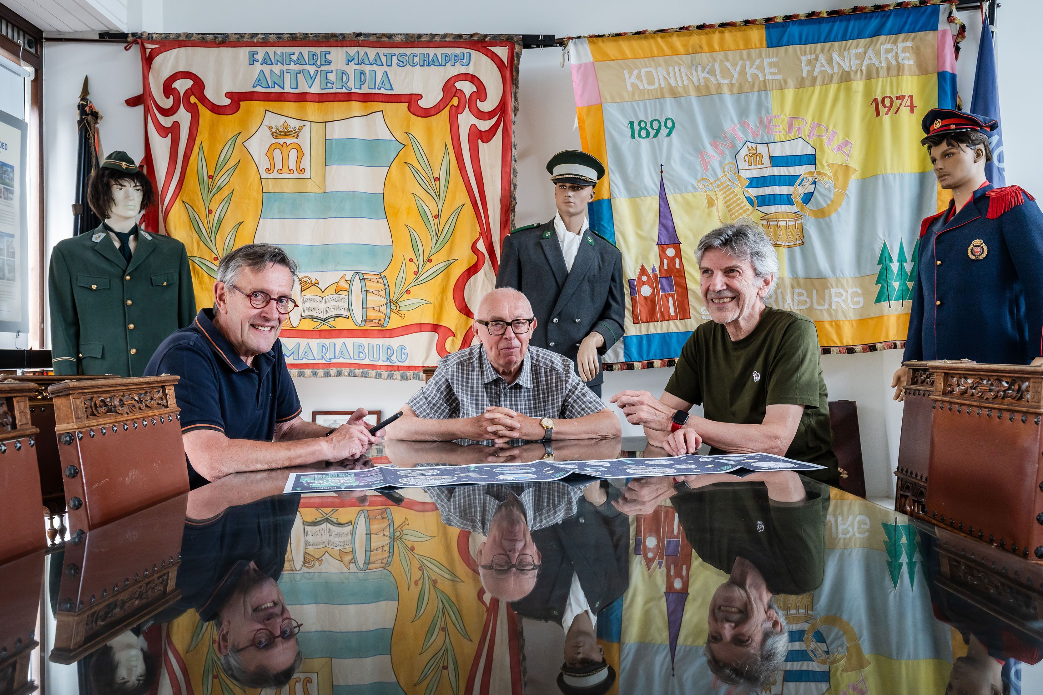 Drie mannen van middelbare leeftijd zitten aan een tafel en kijken lachend naar de camera, op de achtergrond staan drie mannequins in uniform en hangen wapenschildvragen op.