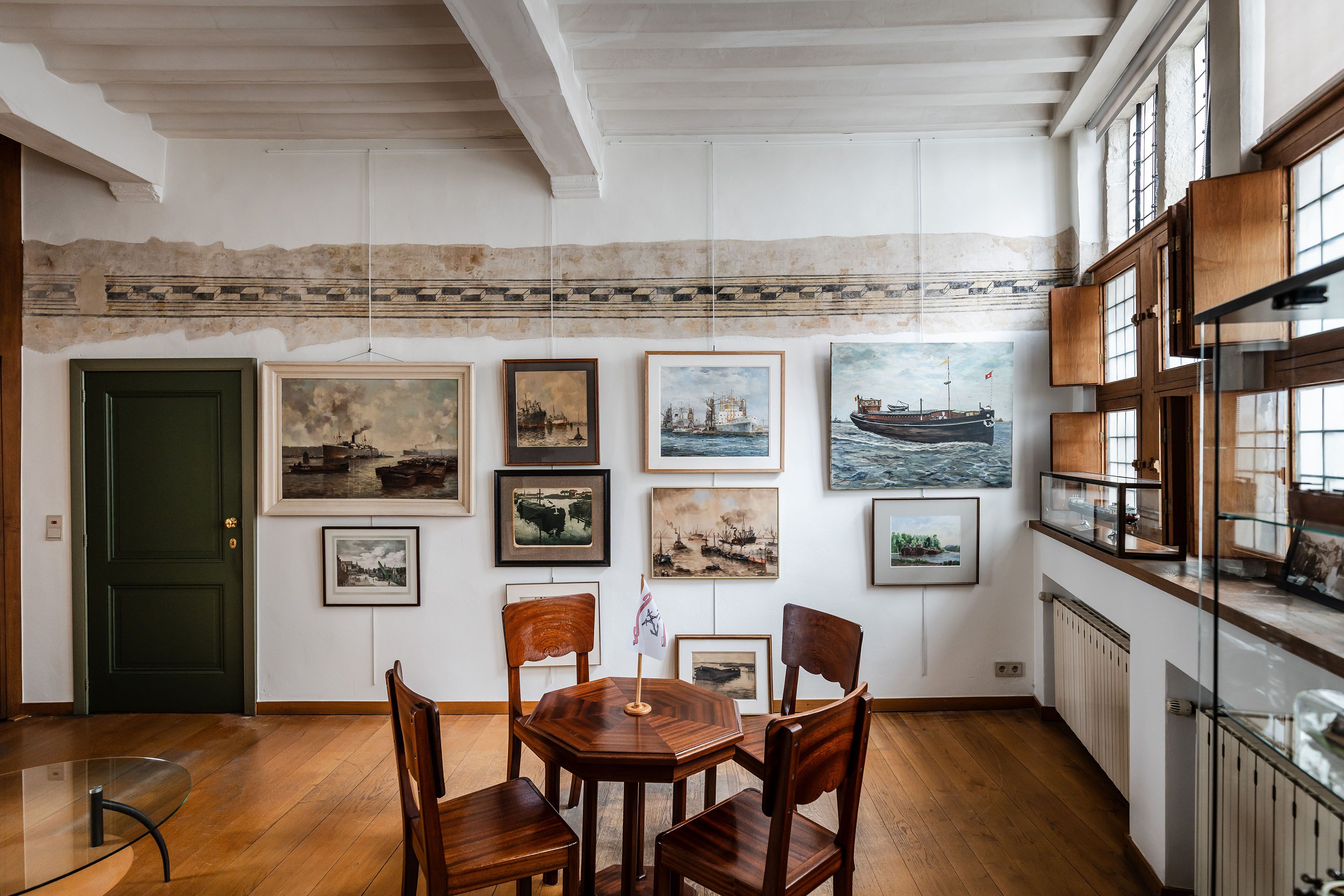 Een witte muur met ingekaderde schilderijen van schepen en ervoor een kleine ronde houten tafel met 4 stoelen.