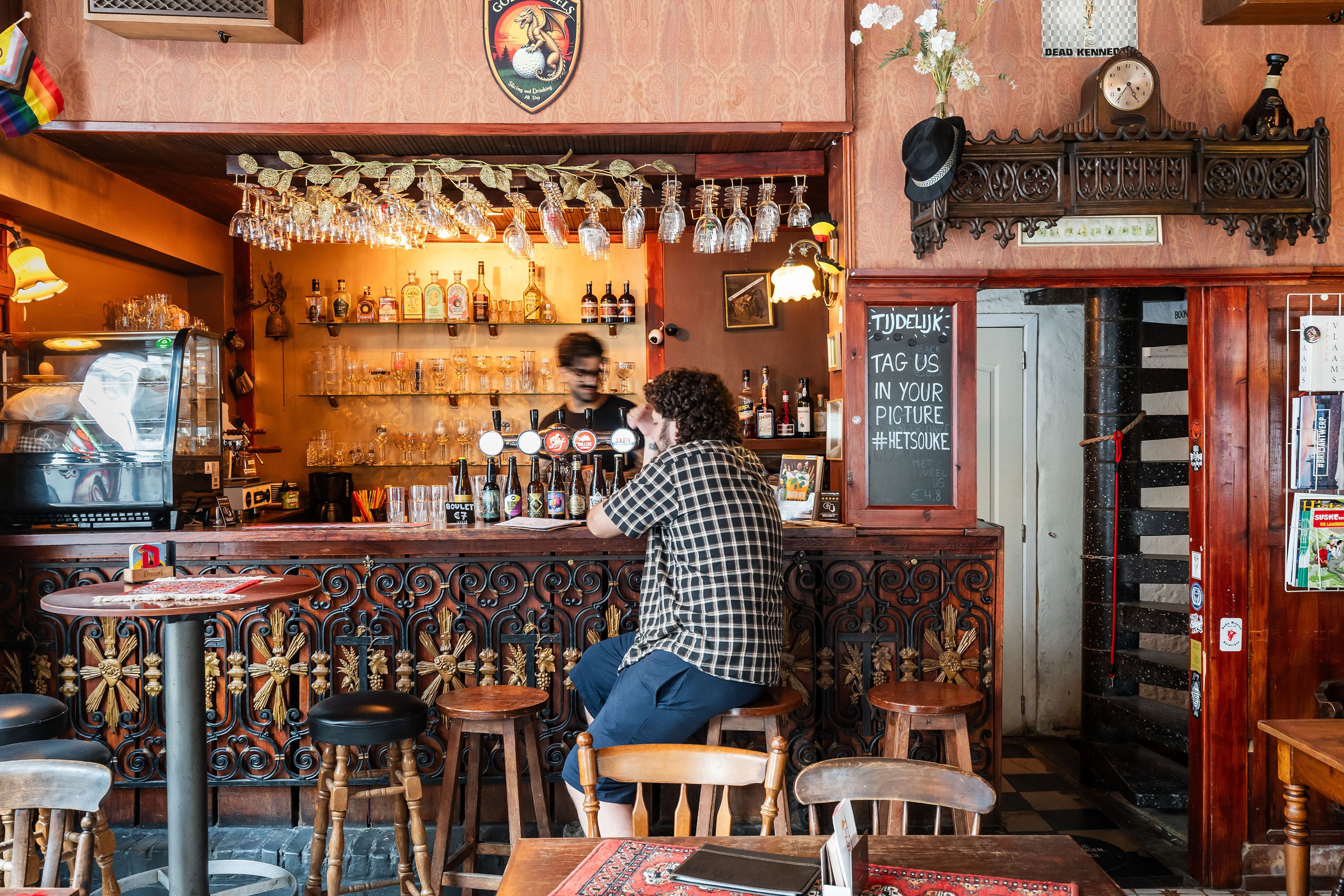 Man zit op barkruk in volkscafé met houten interieur aan de toog. Achter de toog tapt een man een pintje.