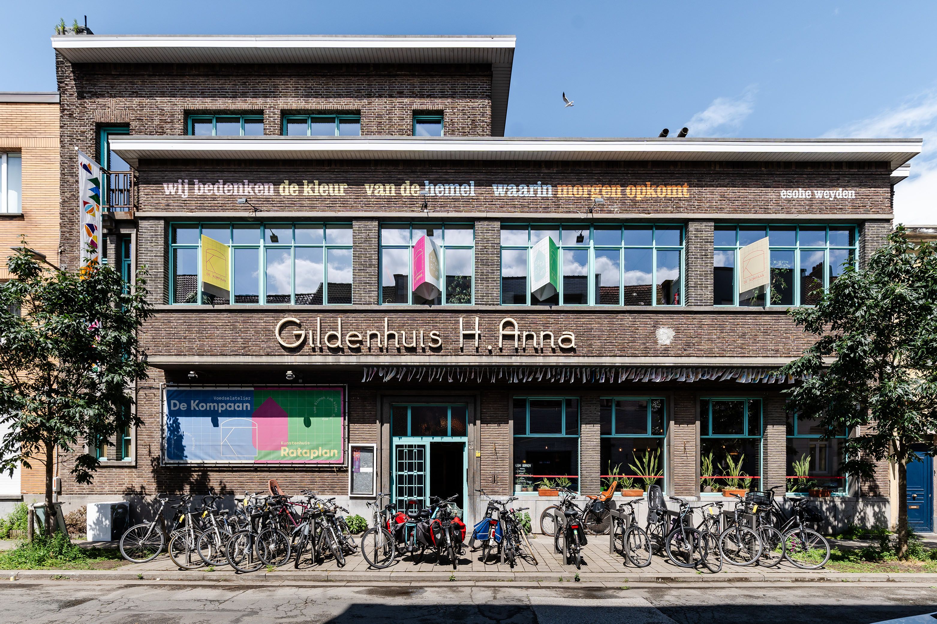 Vooraanzicht van een meerlagig gebouw met een brede gevel. Voor het gebouw staan fietsen geparkeerd en enkele bomen langs de straat. Op het gelijkvloers zijn kleurrijke raampartijen zichtbaar.