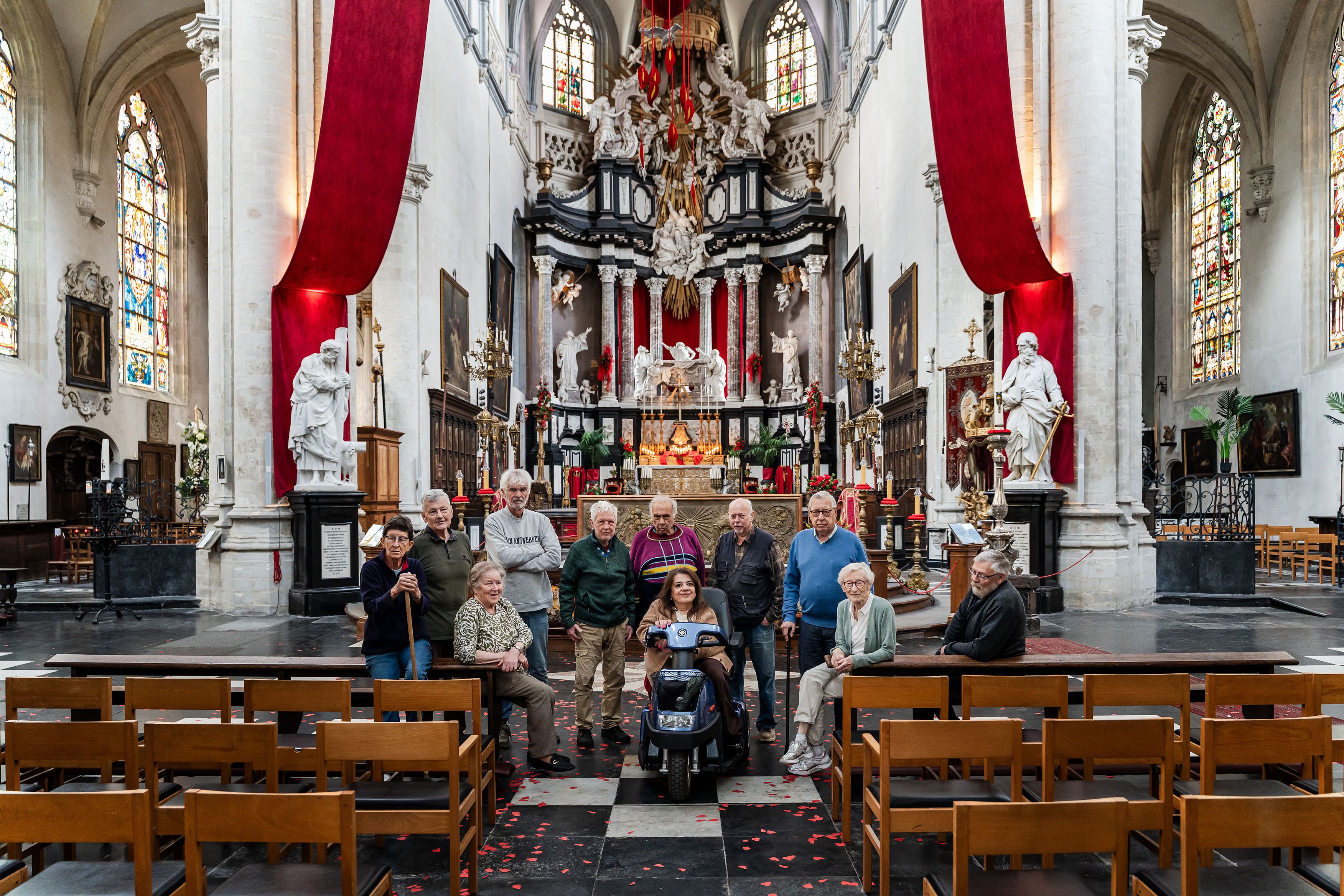 Groepsfoto van een twintigtal personen in het middenpad van een grote kerk. Op de achtergrond staat een rijk versierd altaar met hoge rode doeken aan weerszijden. Op de voorgrond zijn houten kerkbanken zichtbaar. De foto is genomen vanuit de achterkant van de kerk, waardoor zowel de groep als het interieur van het kerkgebouw in beeld zijn.