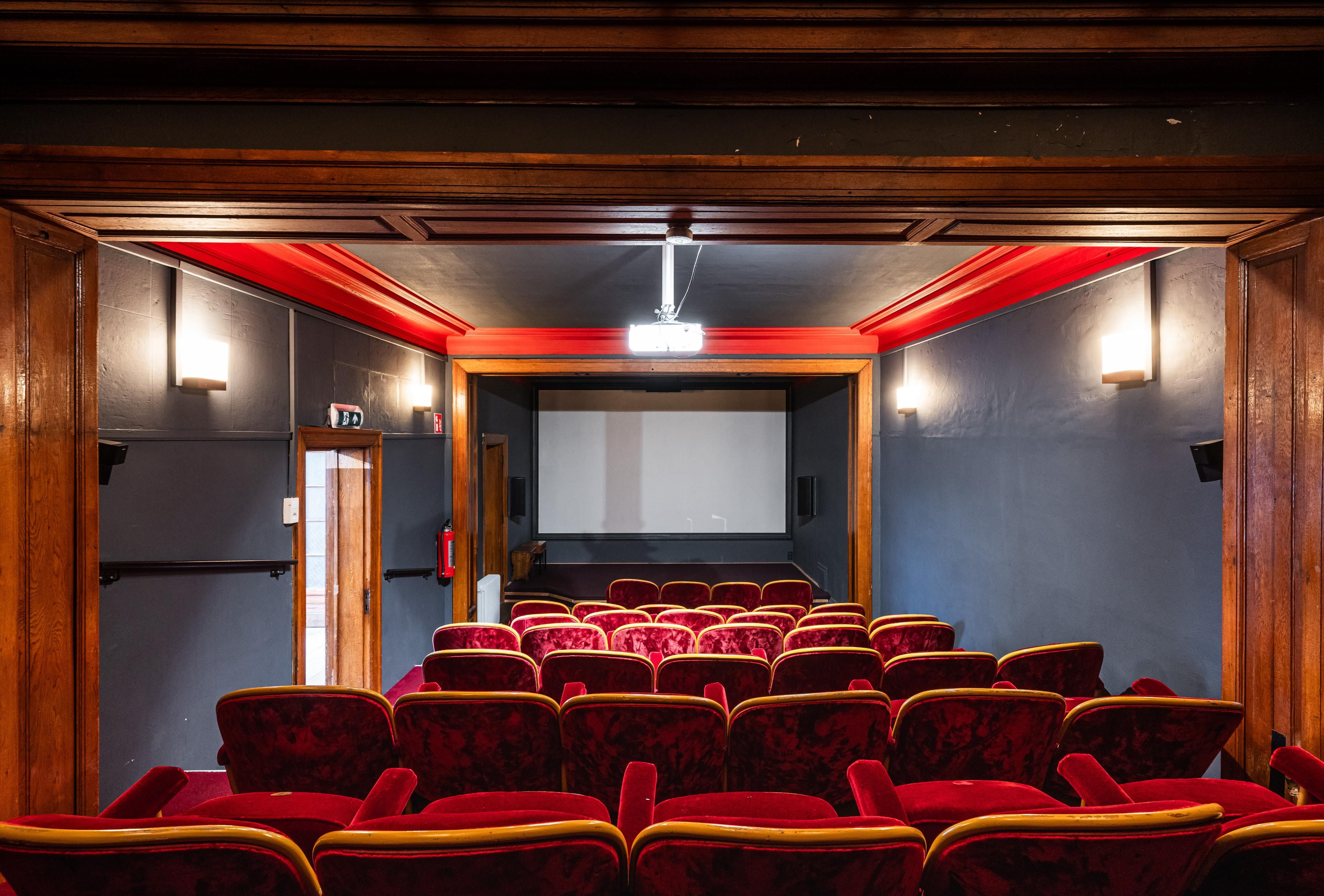 Een zicht vanaf de achterzijde van de filmzaal naar het scherm. De muren en het plafond zijn geschilderd in een grijs-blauw. De rijen stoffen zetels zijn rood.