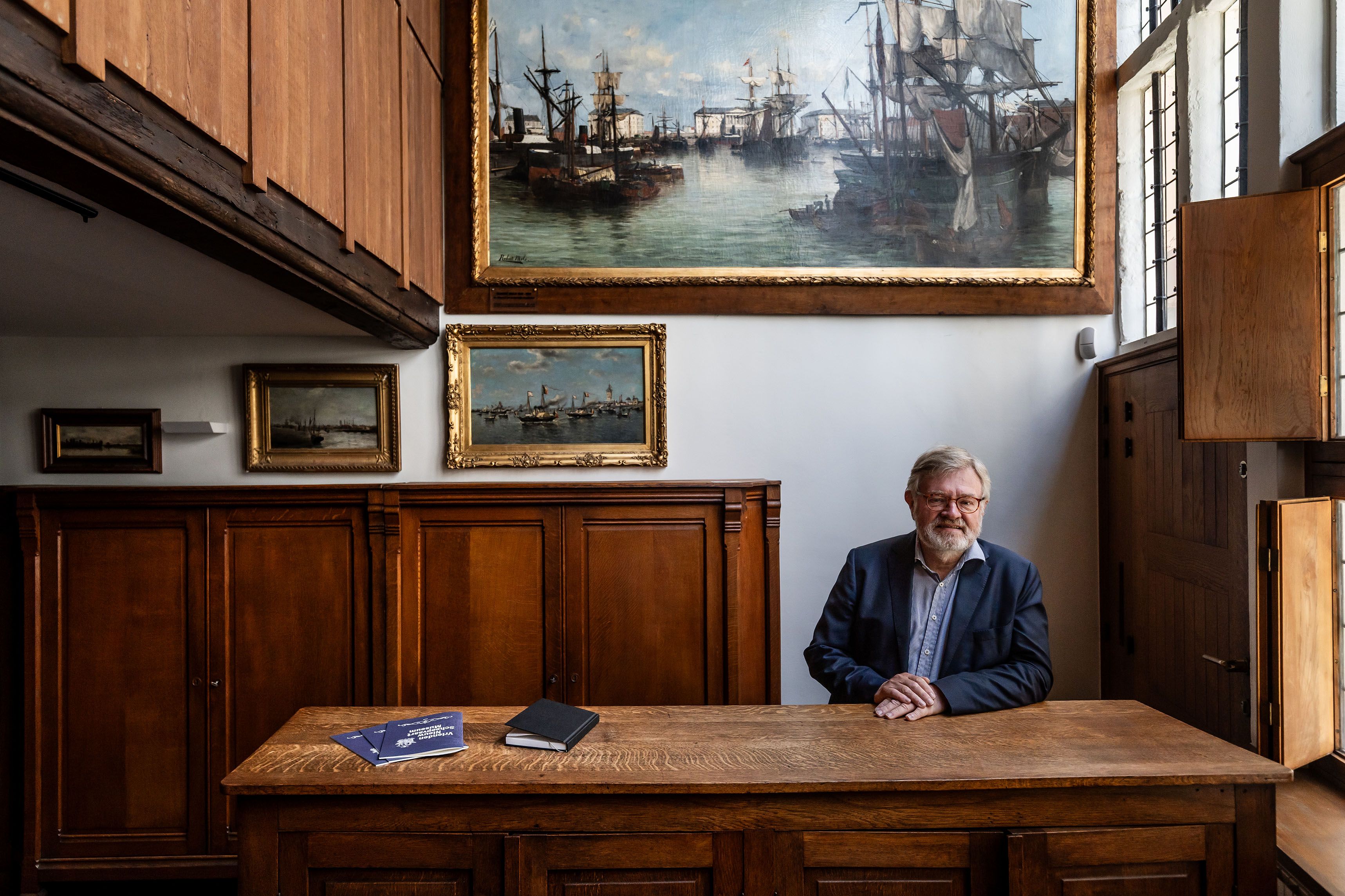 Man van middelbare leeftijd aan een houten bureau met op de achtergrond een schilderij van schepen en een houten kast.