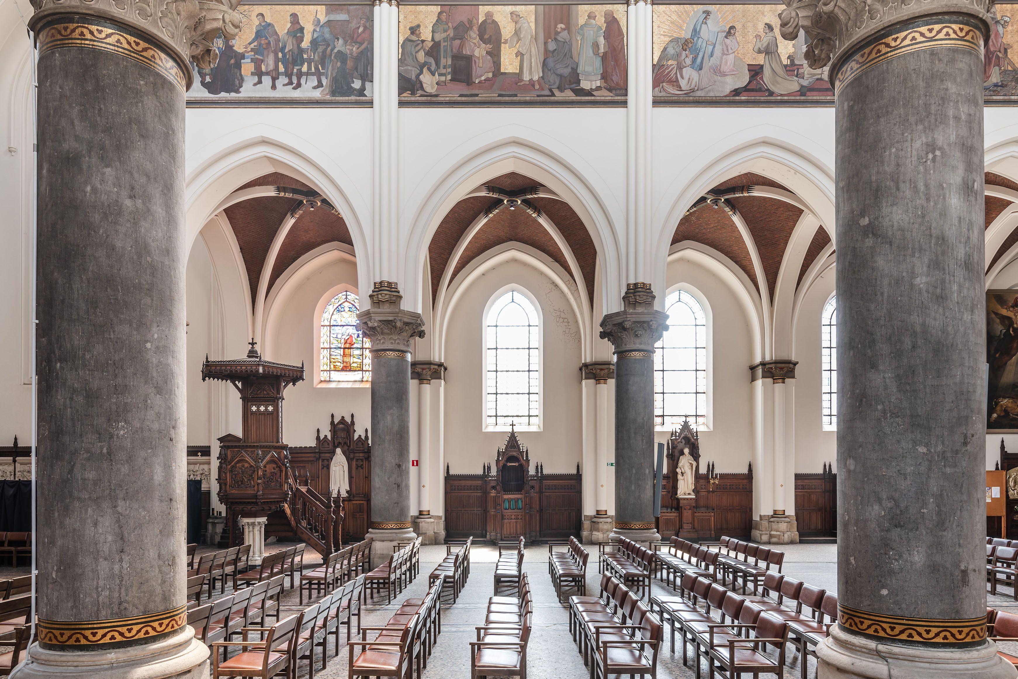 Interieur Sint-Norbertuskerk met grijze zuilen, witte muur en gekleurde muurschilderingen en lange rijen stoelen.