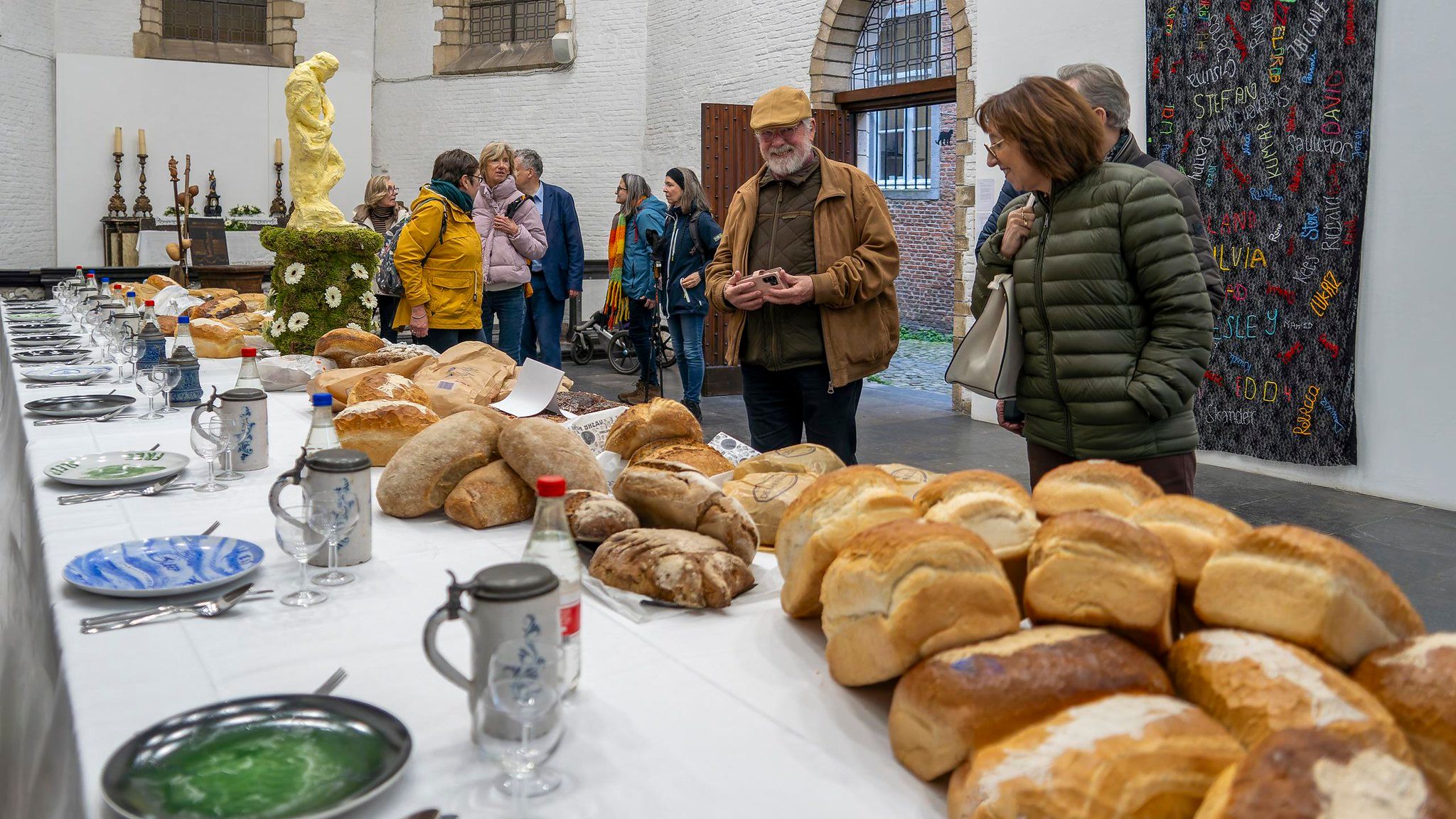 Gedekte pelgrimstafel met broden borden en drinkbekers. Een tiental mensen loopt rond in de kapel waar de tafel staat.