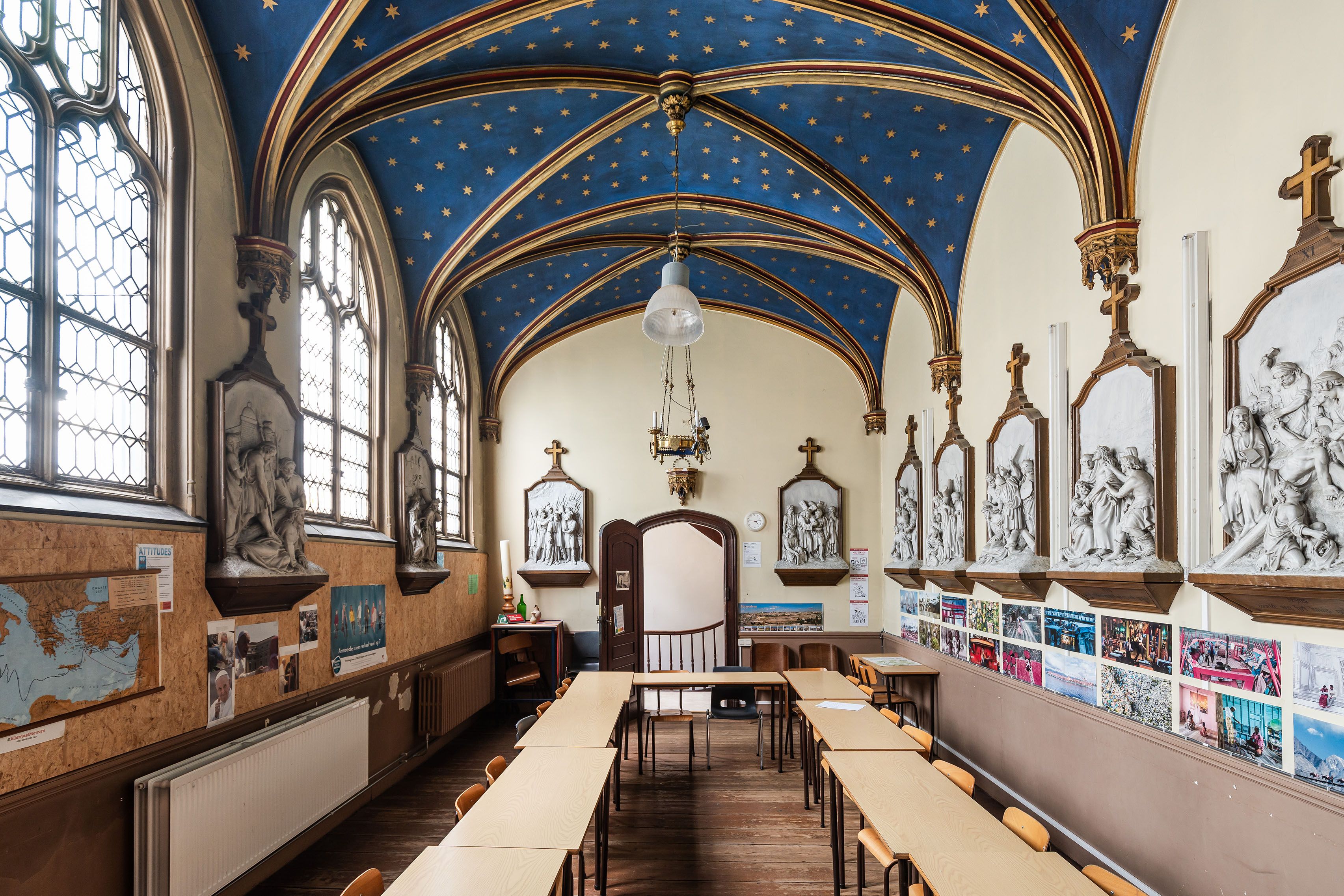 Ruimte in het erfgoedpand van Celst met afgeronde bogen op het helderblauwe plafond. Er staan stoelen en banken in de ruimte met religieuze beelden aan de wanden.