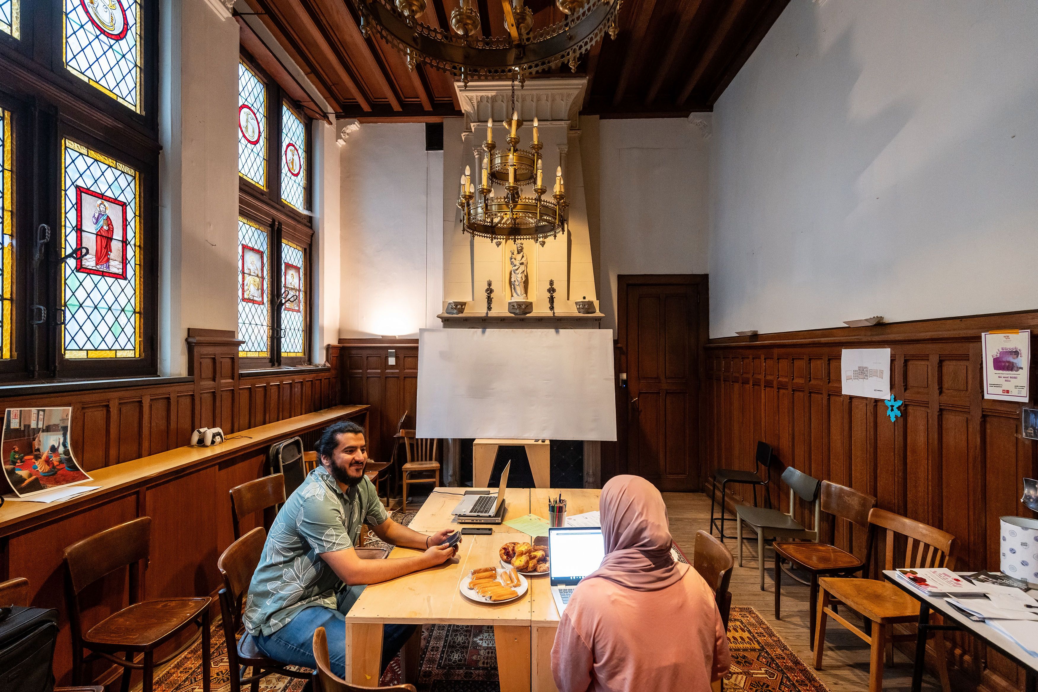 Man en vrouw met hoofddoek praten met elkaar aan een tafel, er staat een bord met koekjes op de tafel.