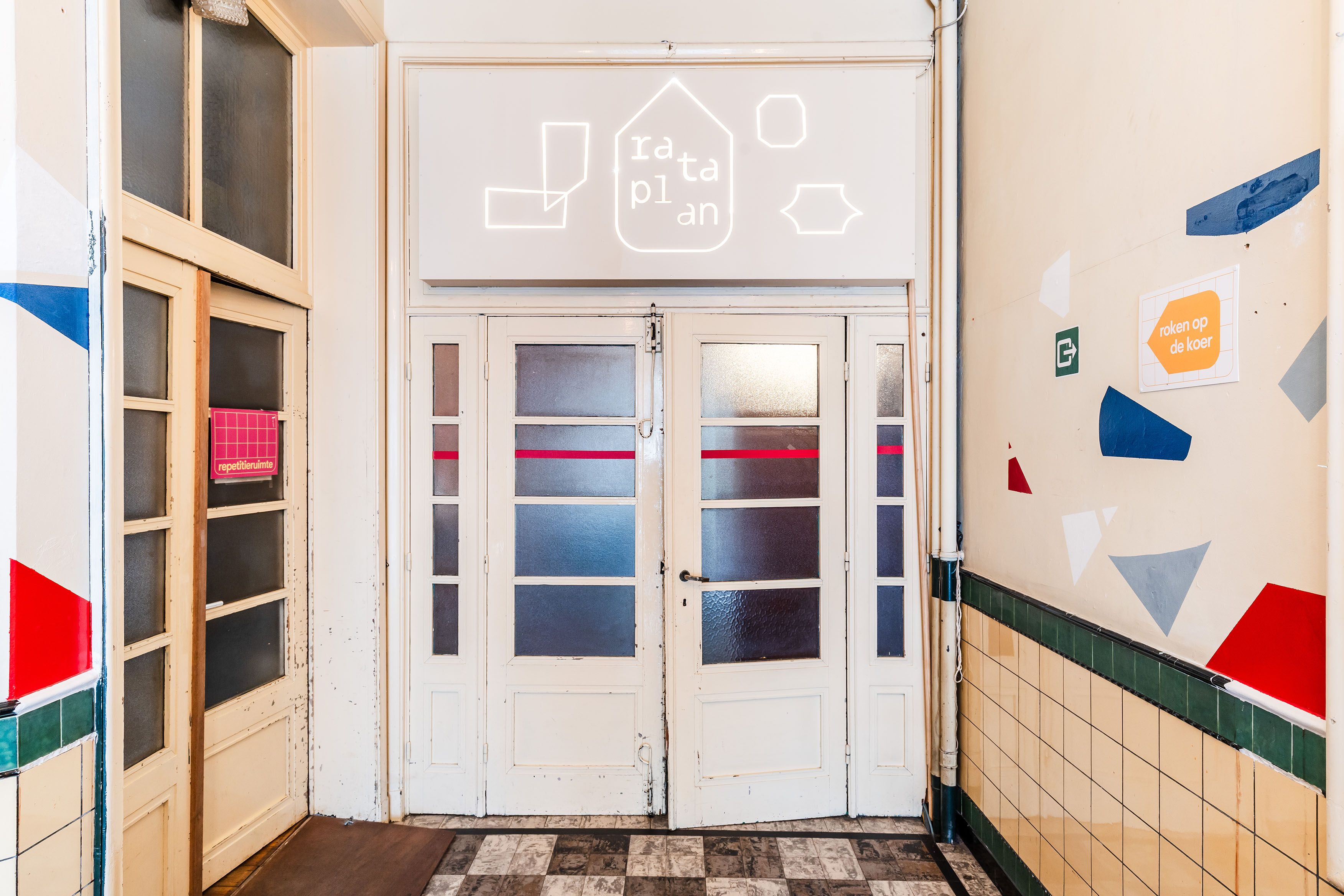 Hal met dubbele glazen deuren, lichte muren en decoratieve gekleurde lijnen op de wanden. Boven de deuren zijn pictogrammen aangebracht en aan weerszijden hangen bewegwijzeringsborden.