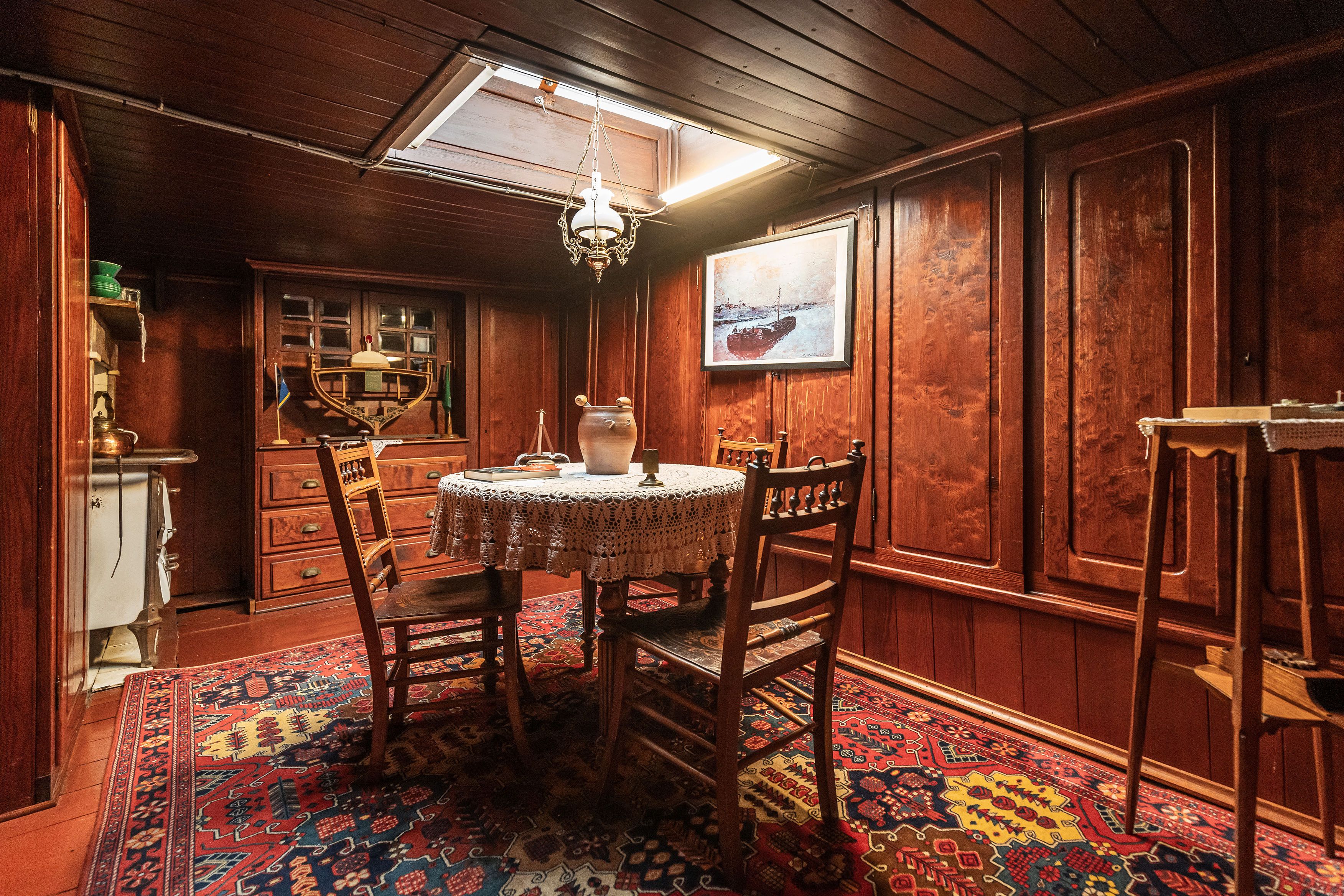 Houten kajuit met in het midden een tafel met kanten tafelkleed en 2 stoelen op een tapijt met rood blauw en geel