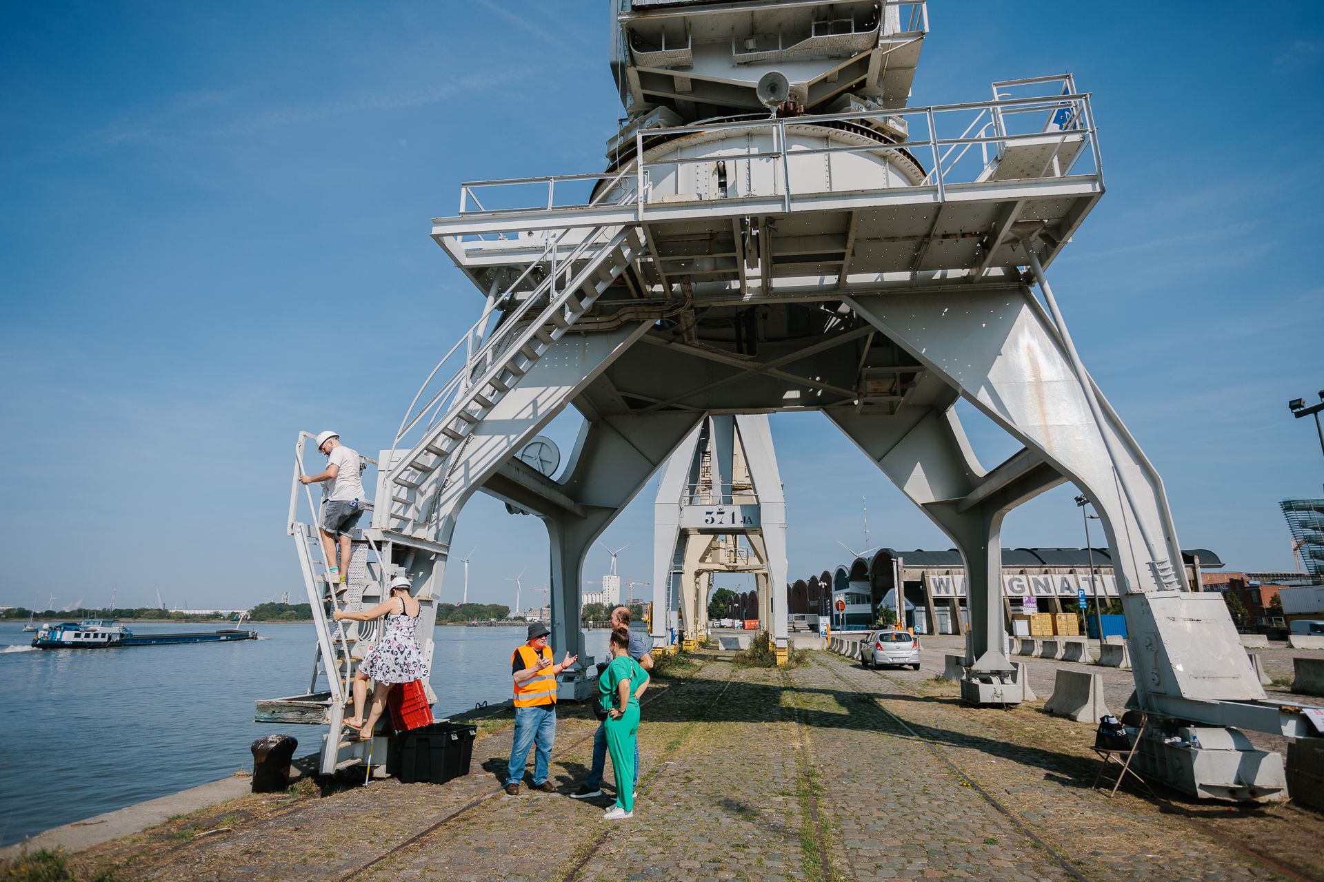 Detail van het onderstel van één van de havenkranen. Een vrijwilliger geeft op de grond uitleg aan twee bezoekers. Twee andere bezoekers beklimmen de ladder voor een bezoek aan de havenkraan.