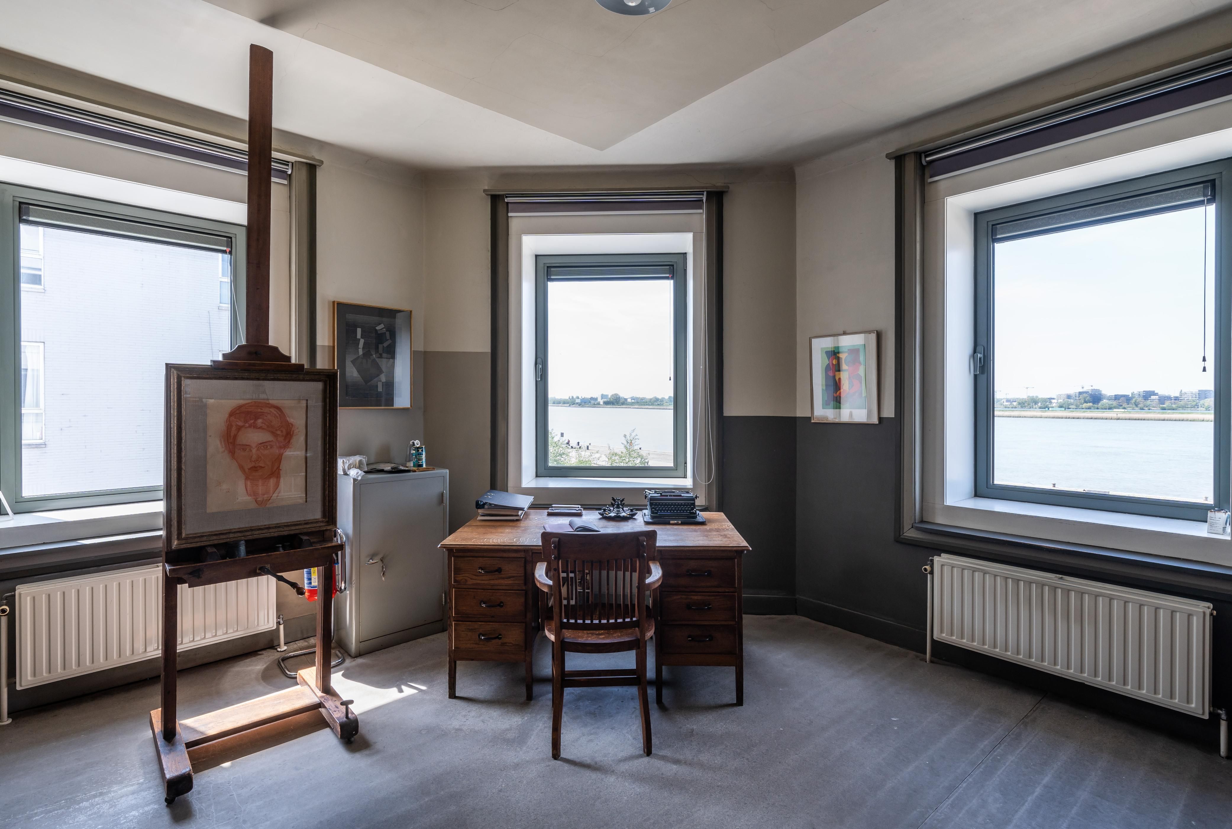 Atelier met bureau en schildersezel en muren in tinten grijs en beige.
