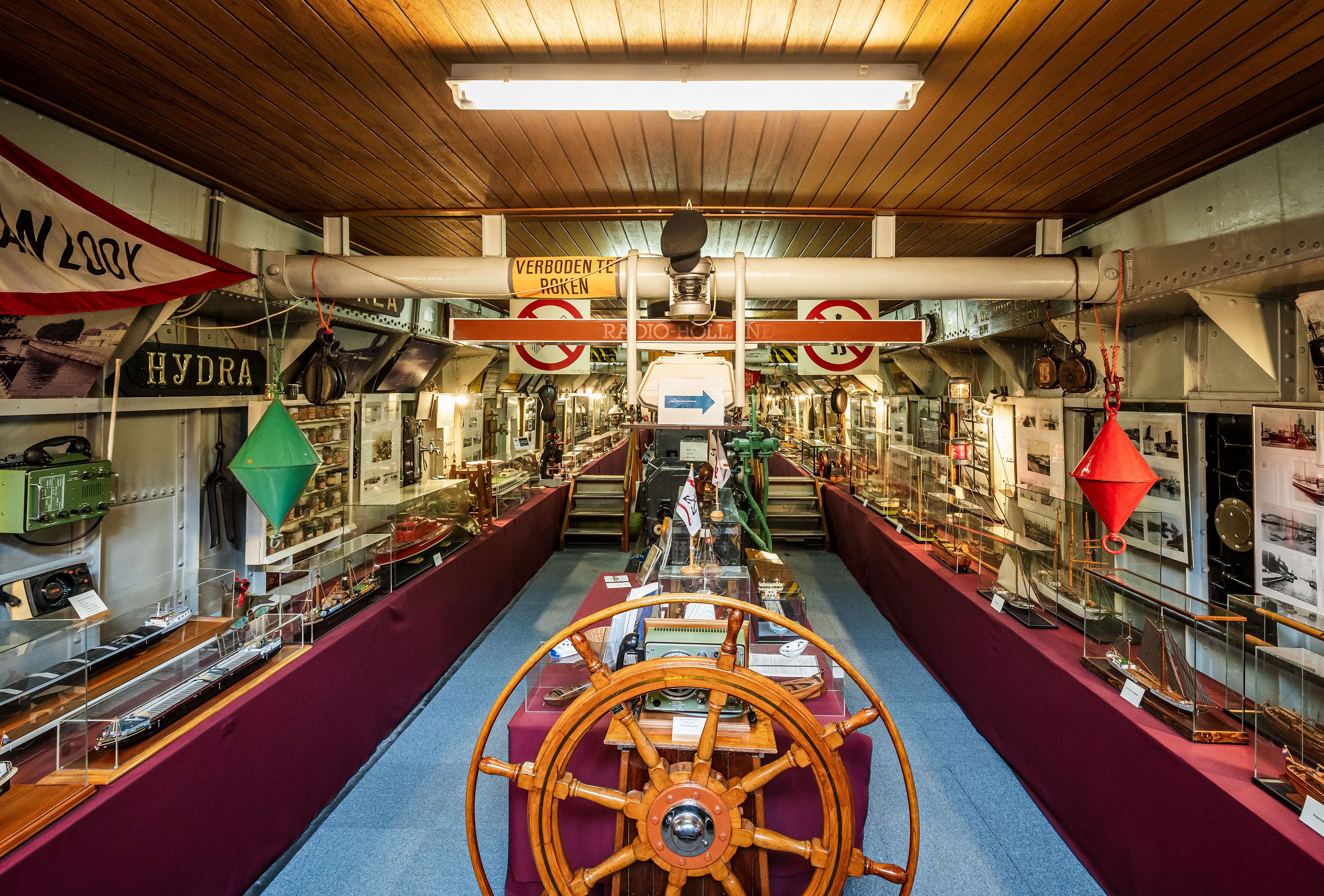 Museumruimte in schip met een roer en veel foto's en schaalmodellen van schepen