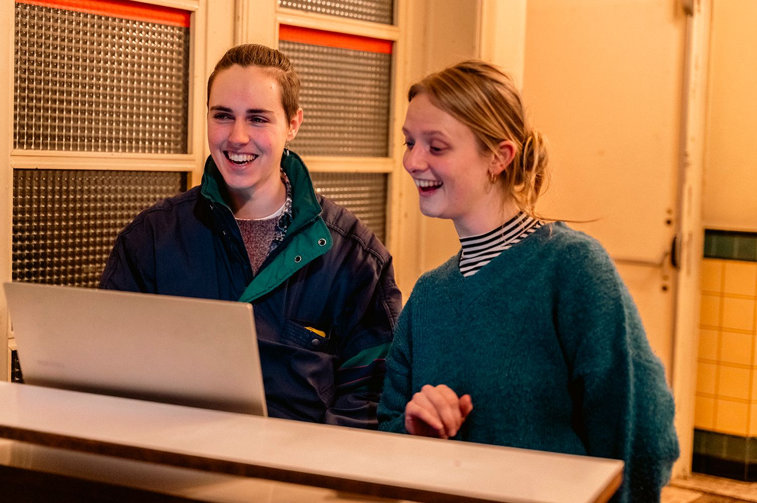 Twee personen staan achter een balie of werkplek en kijken naar een computerscherm. De ruimte is warm verlicht en ingericht als onthaal- of ontvangstomgeving.
