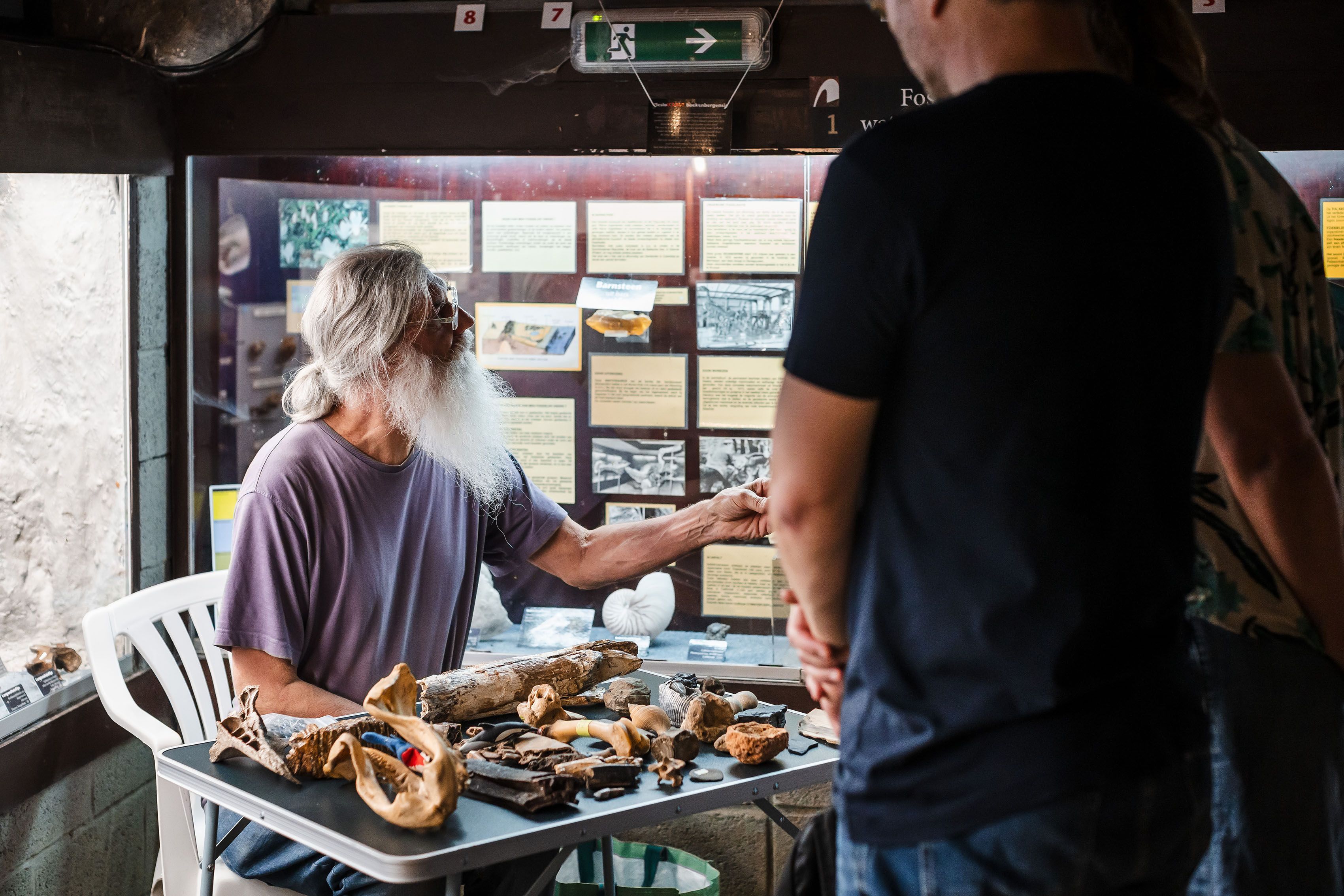 Een vrijwilliger toont en vertelt over museumstukken. Hij zit aan een tafeltje met daarop museumobjecten.