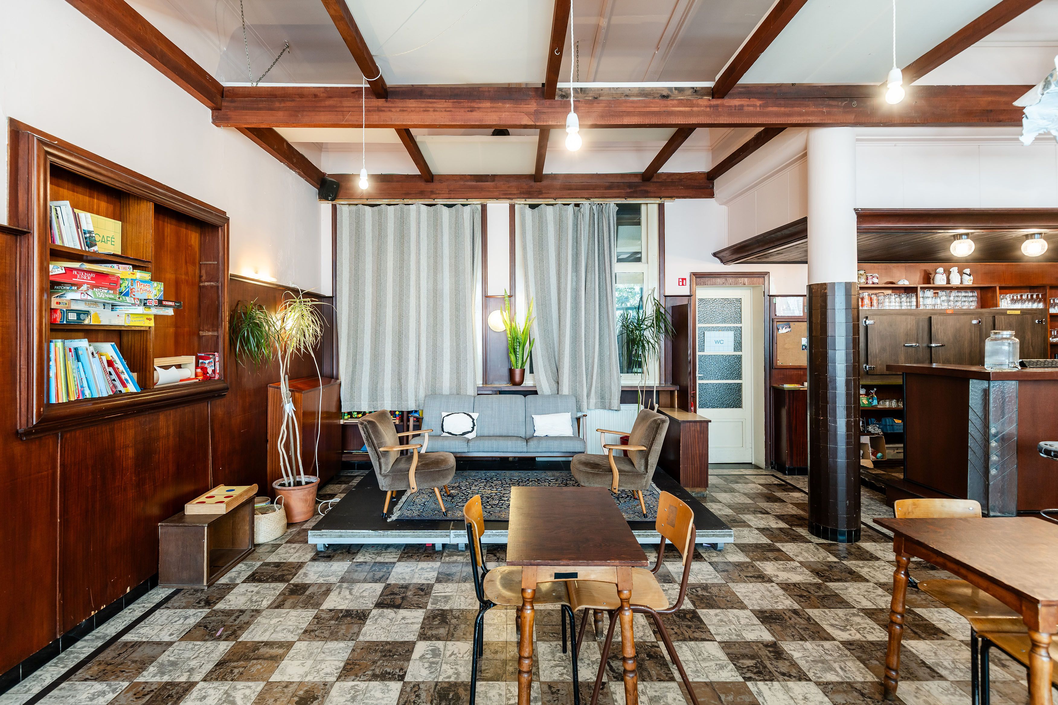 Gezellige ruimte met zwart-bruine tegelvloer, houten wandafwerking en een rechthoekige tafel met stoelen. Grote ramen laten daglicht binnen. In de ruimte staan planten, kasten en decoratieve elementen.