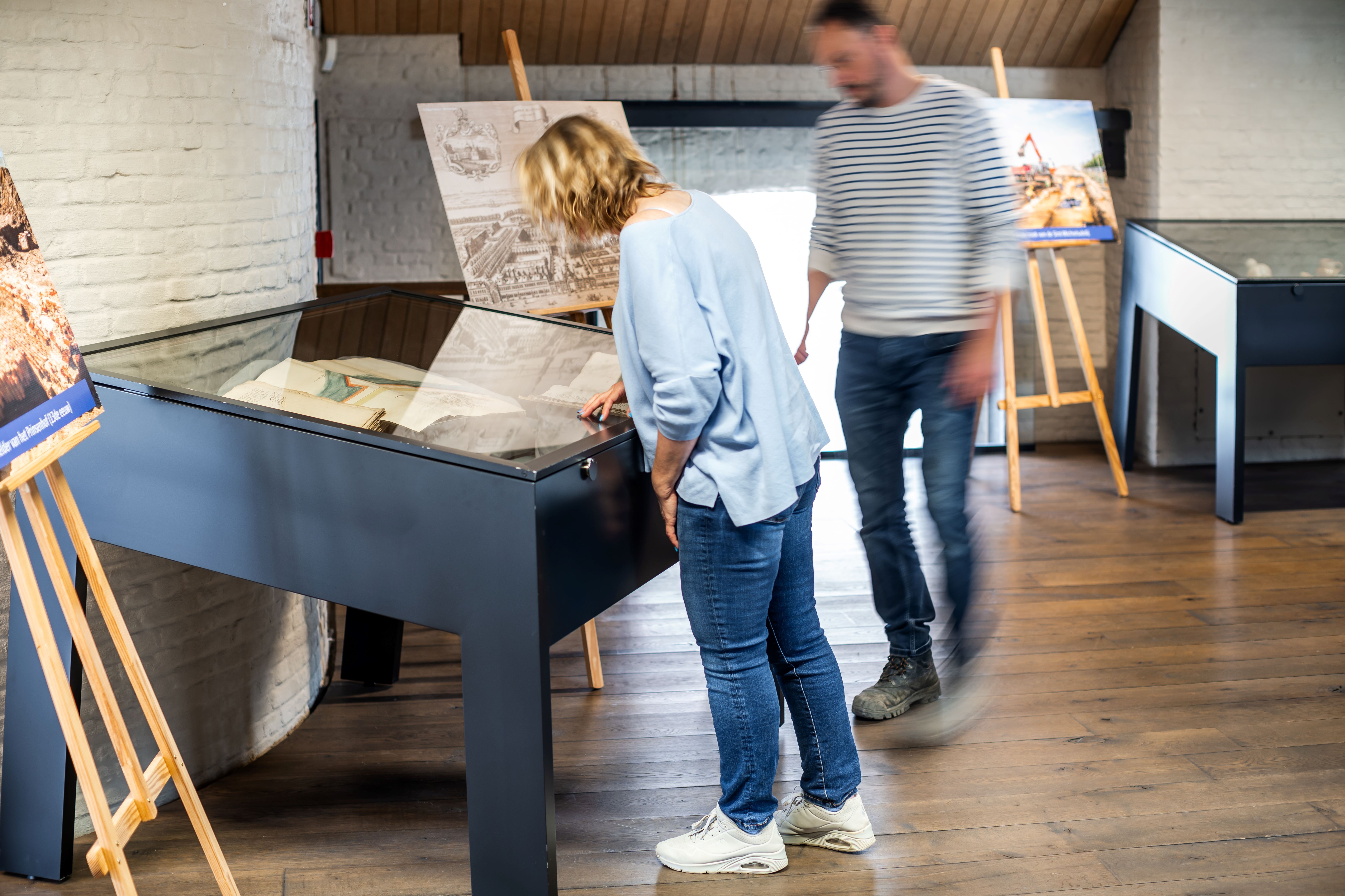 Bezoekers bekijken een archeologische expo op de vierde verdieping. Ze buigen over een glazen kast waar oude kaarten in liggen.