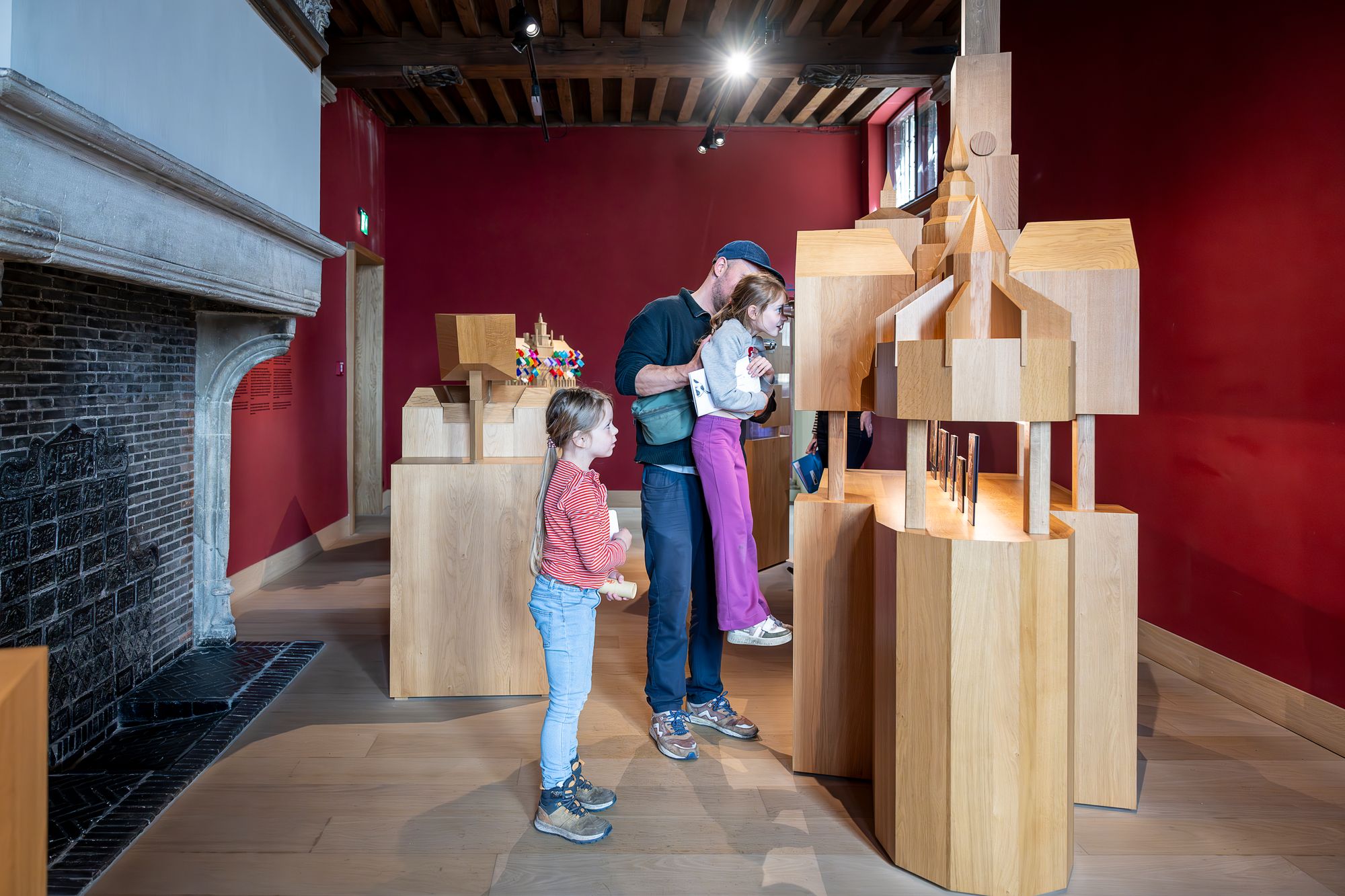 Een man en 2 kinderen, meisjes zoeken in een tentoonstelling naar hints voor en zoektocht. De man tilt een van de kinderen op om beter te kunnen zien.