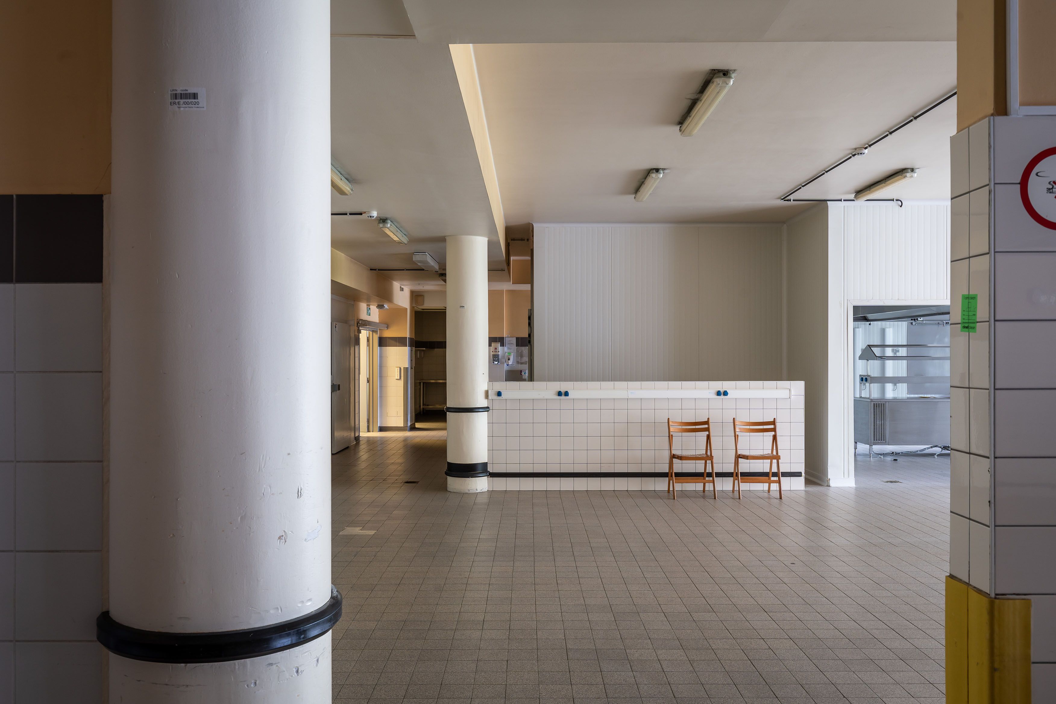 Lege ruimte Sint-Erasmusziekenhuis. 2 witte zuilen en kleine witte vierkante vloertegels en 2 houten klapstoelen.