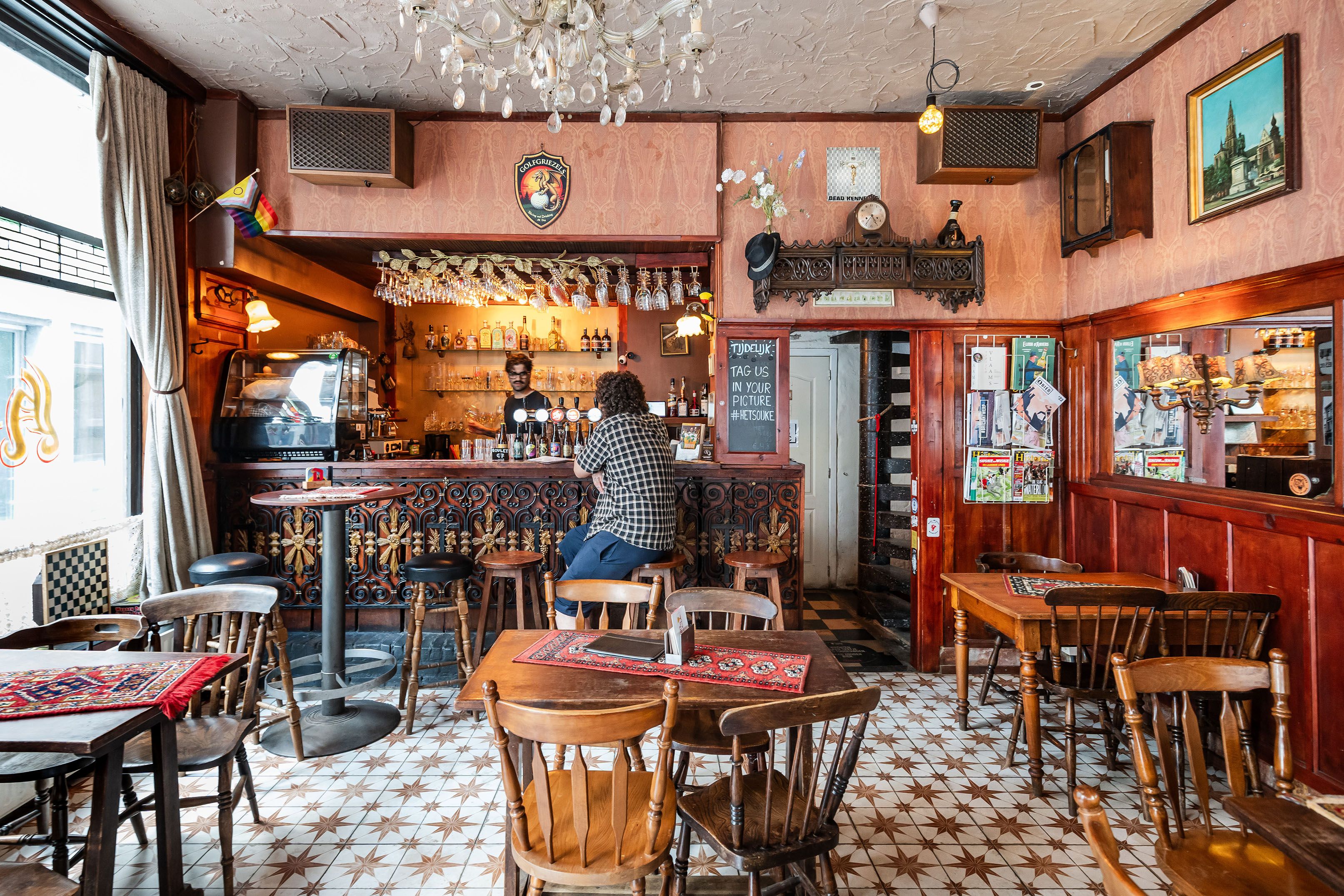 Man zit op barkruk in volkscafé met houten interieur aan de toog. Achter de toog tapt een man een pintje.