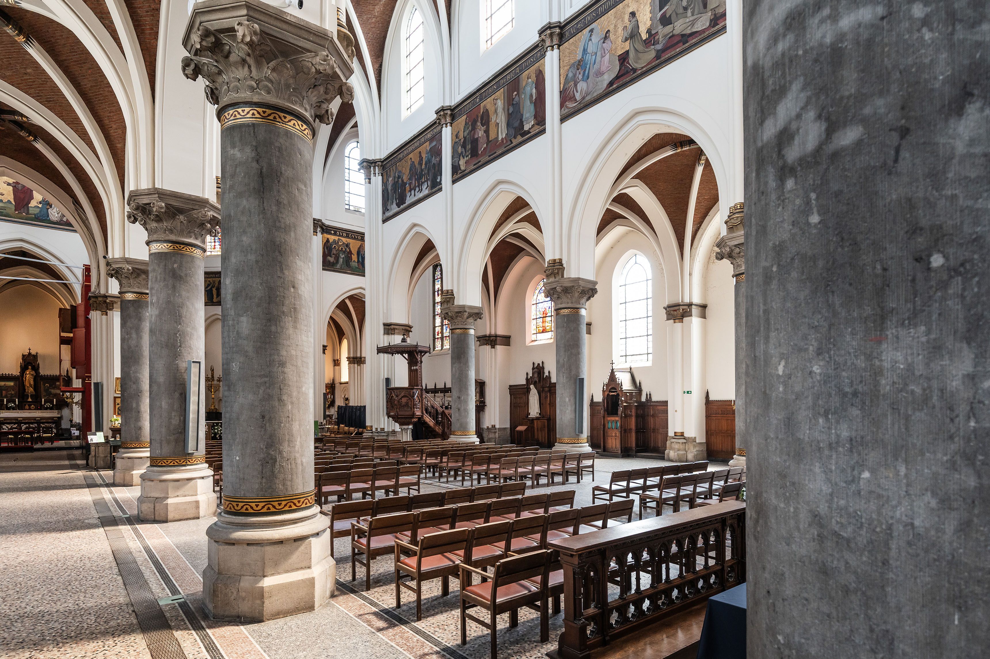 Interieur Sint-Norbertuskerk met grijze zuilen, witte muur en muurschildering in bandvorm.