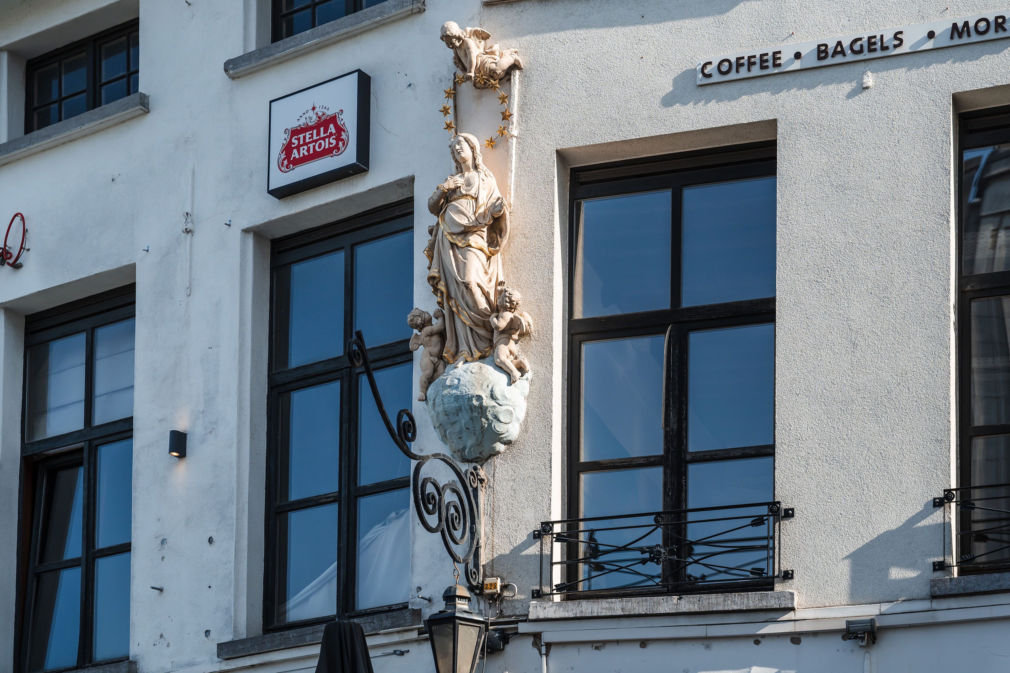 Witgeschilderde madonna met gouden details aan de rand van haar kleding. Een engel boven haar houdt een sterrenkrans boven haar hoofd. Onderaan kijken twee biddende engelen naar haar op. De lamp van de lantaarn onder de sokkel is verdwenen.