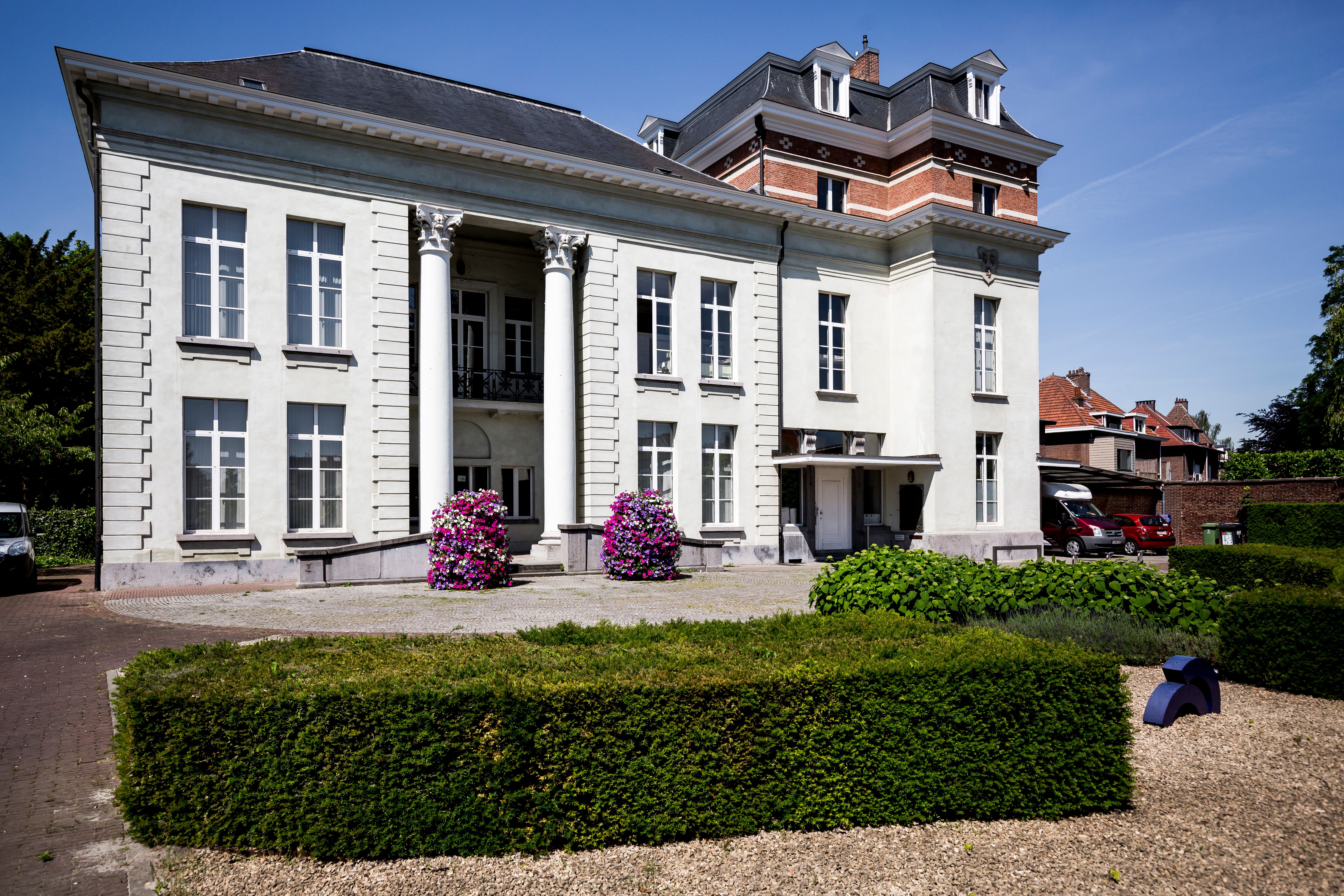 Wit geschilderd kasteel met ervoor 2 bloempotten met roze bloemen en een lage groene haag.