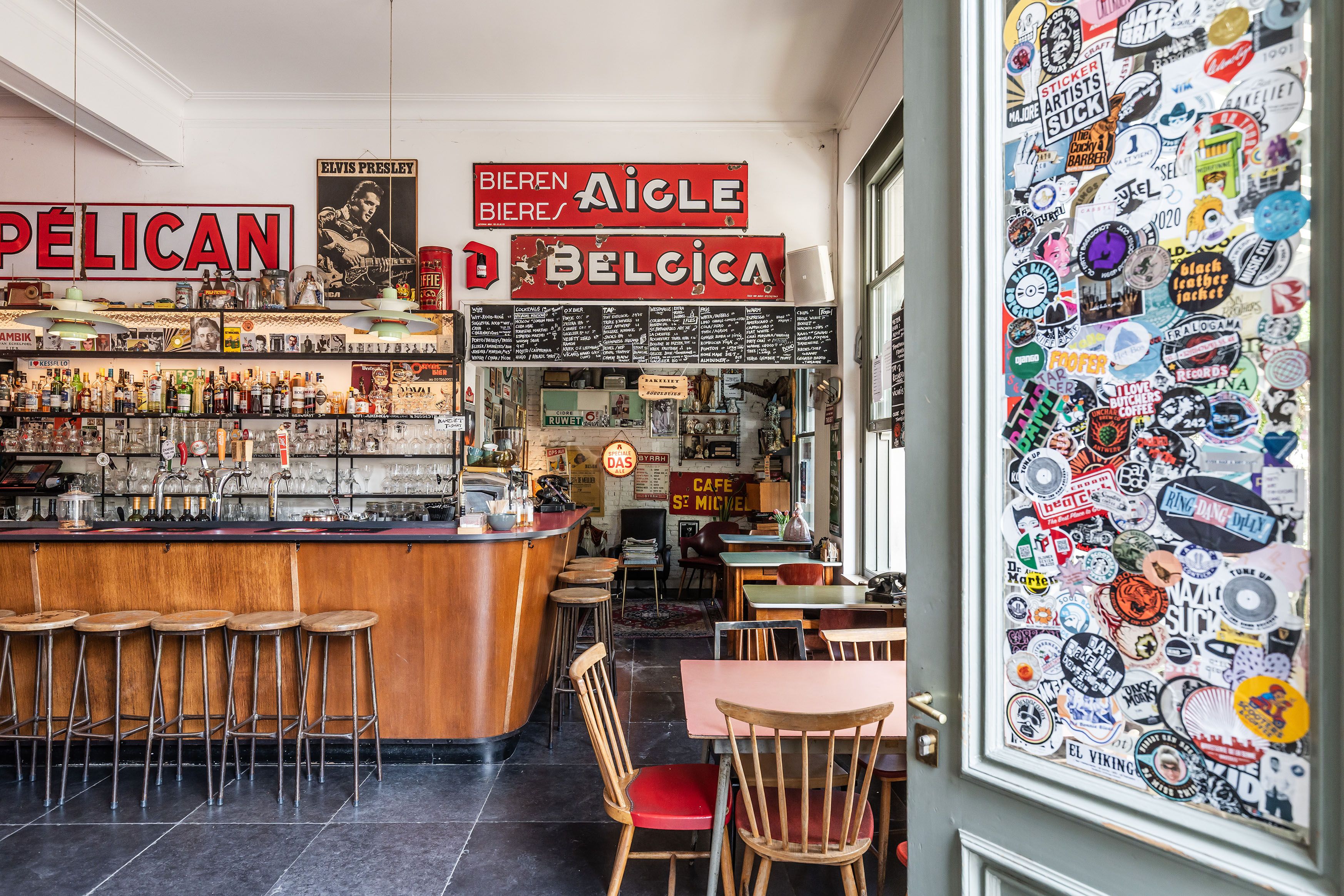 Interieur Bar Bakeliet met oude metalen logo's aan de muur, een houten toog met krukken en veel stickers op de deur.