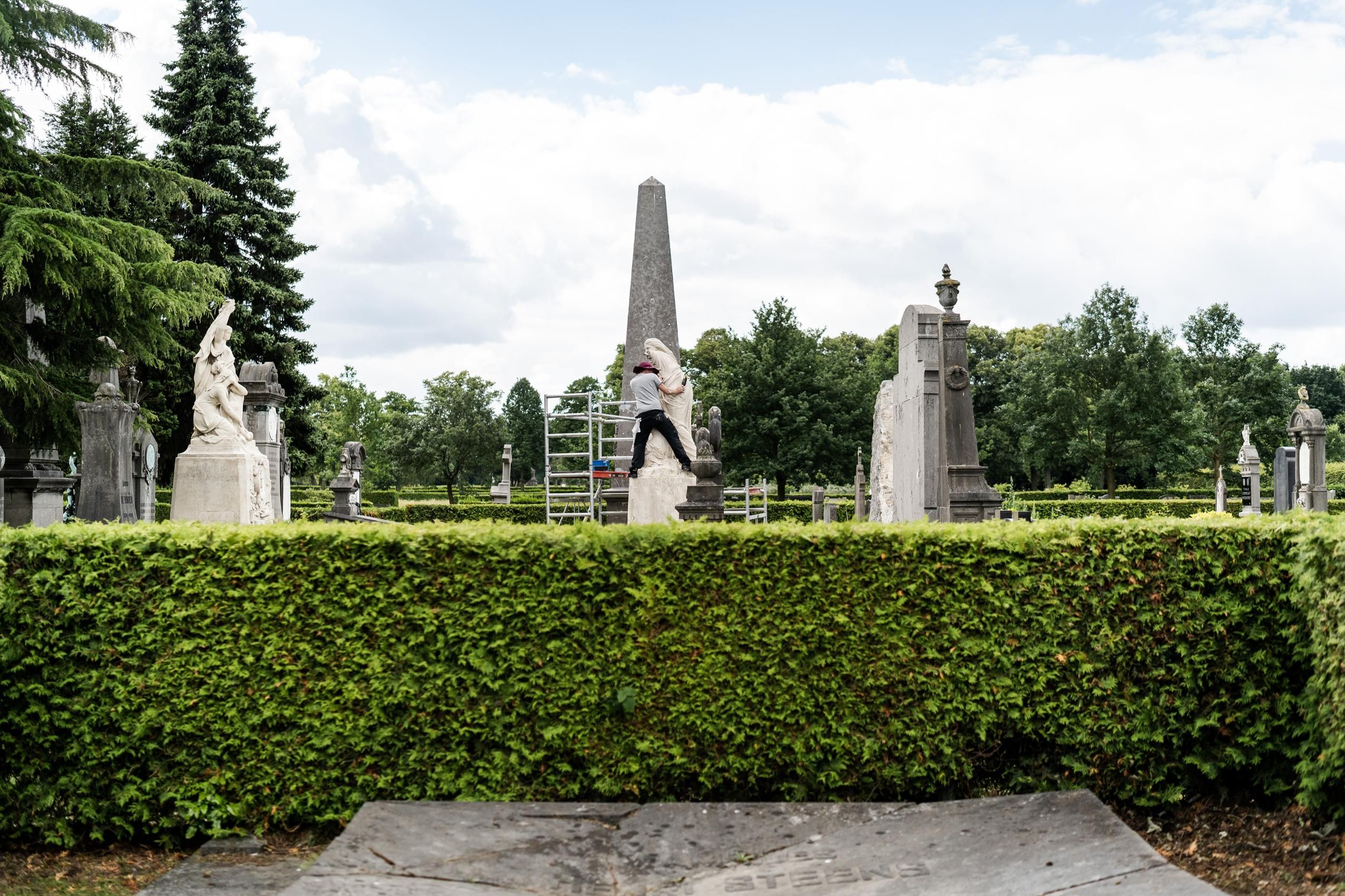 Op de voorgrond bevindt zich een haag. Achter de haag staan monumentale grafmonumenten. Ventraal wordt één van de grafmonumenten gerestaureerd.