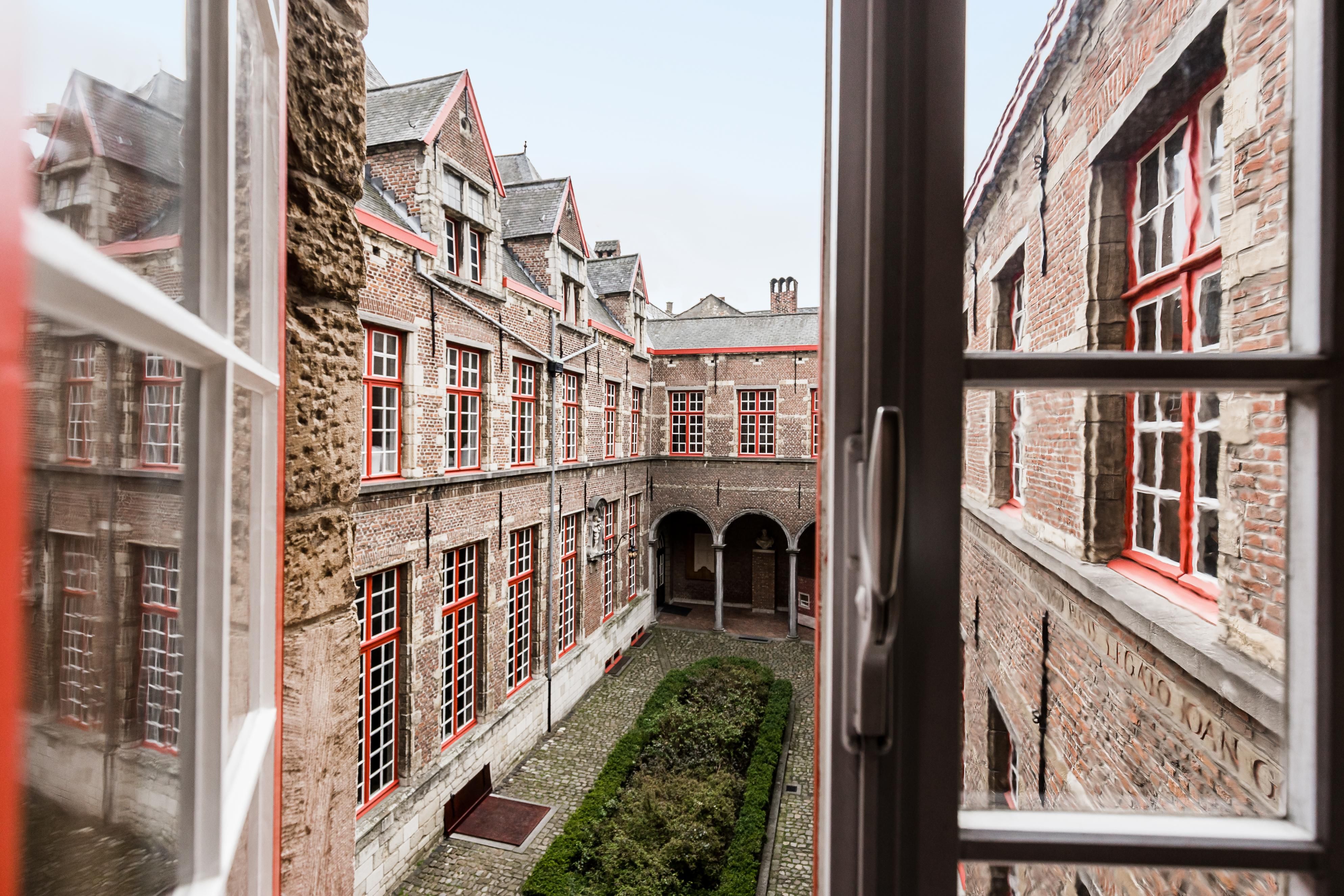 Zicht door een open raam op het binnenplein van het Maagdenhuis. Rode bakstenen muren rond een binnenplein met kasseien en in het midden een lage groene beplanting.