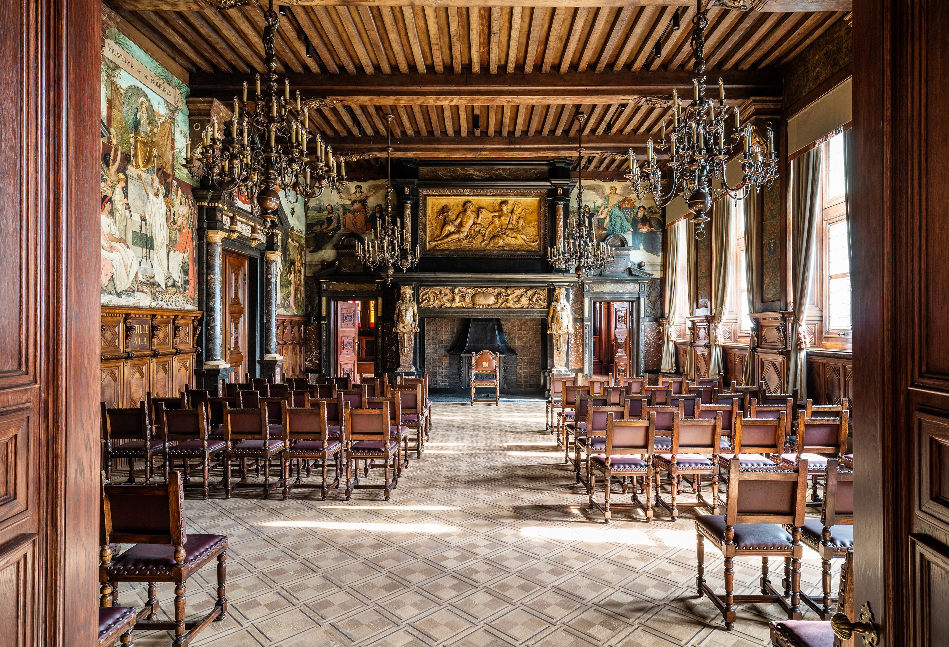 Trouwzaal stadhuis met houten lambrisering en erboven schilderijen, een rijkelijk uitgewerkte schouw in marmer, verschillende rijen stoelen op een parketvloer in geometrisch patroon.