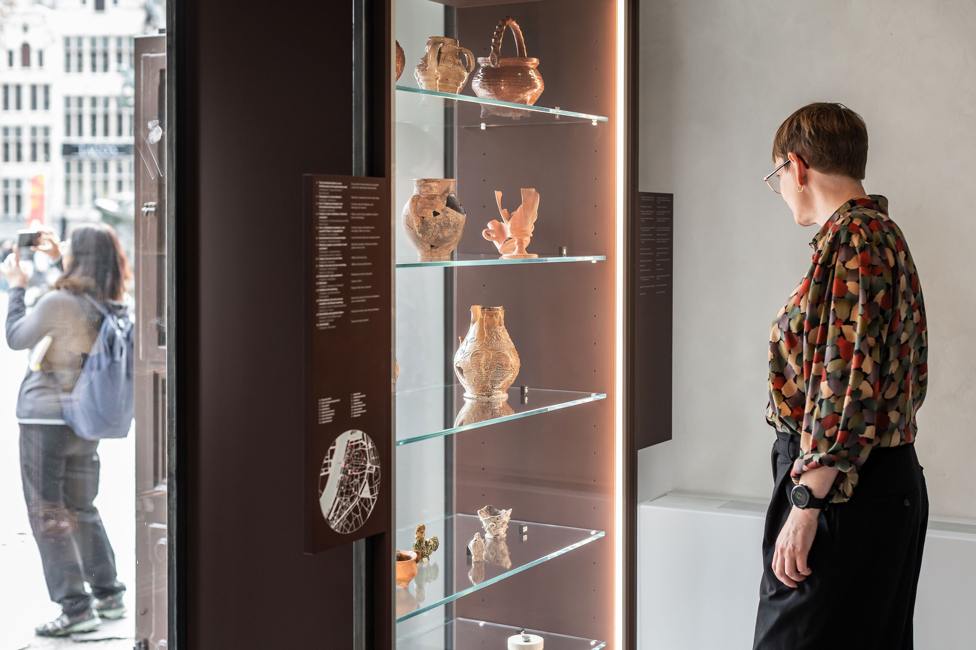 Vrouw kijkt naar archeologische vondsten in glazen kast. Erachter staat een vrouw die een foto neemt.
