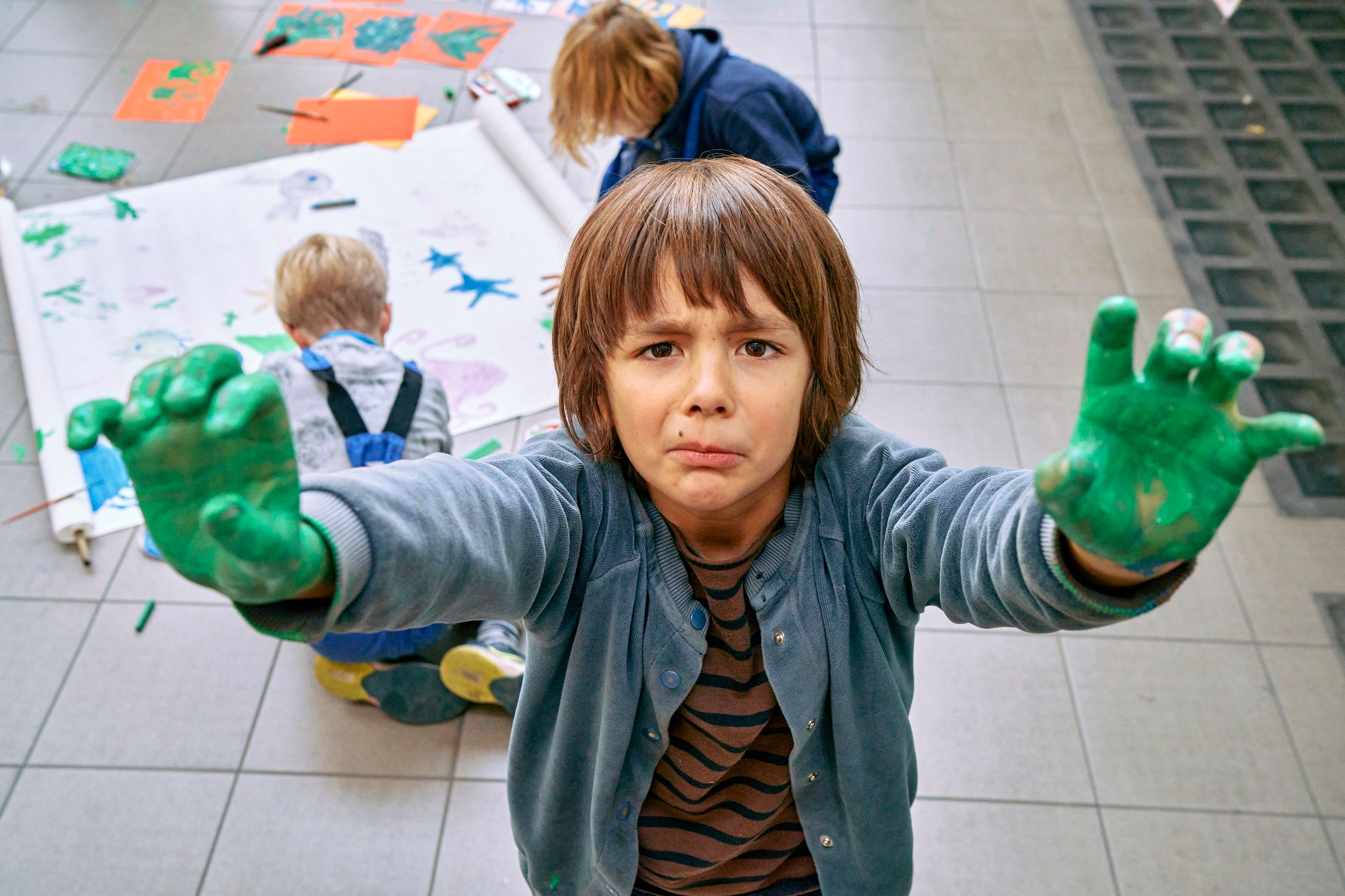 Jongen van ongeveer 6 jaar toont groen geschilderde handen. Achter hem schilderen 2 kinderen een blad dat op de grond ligt