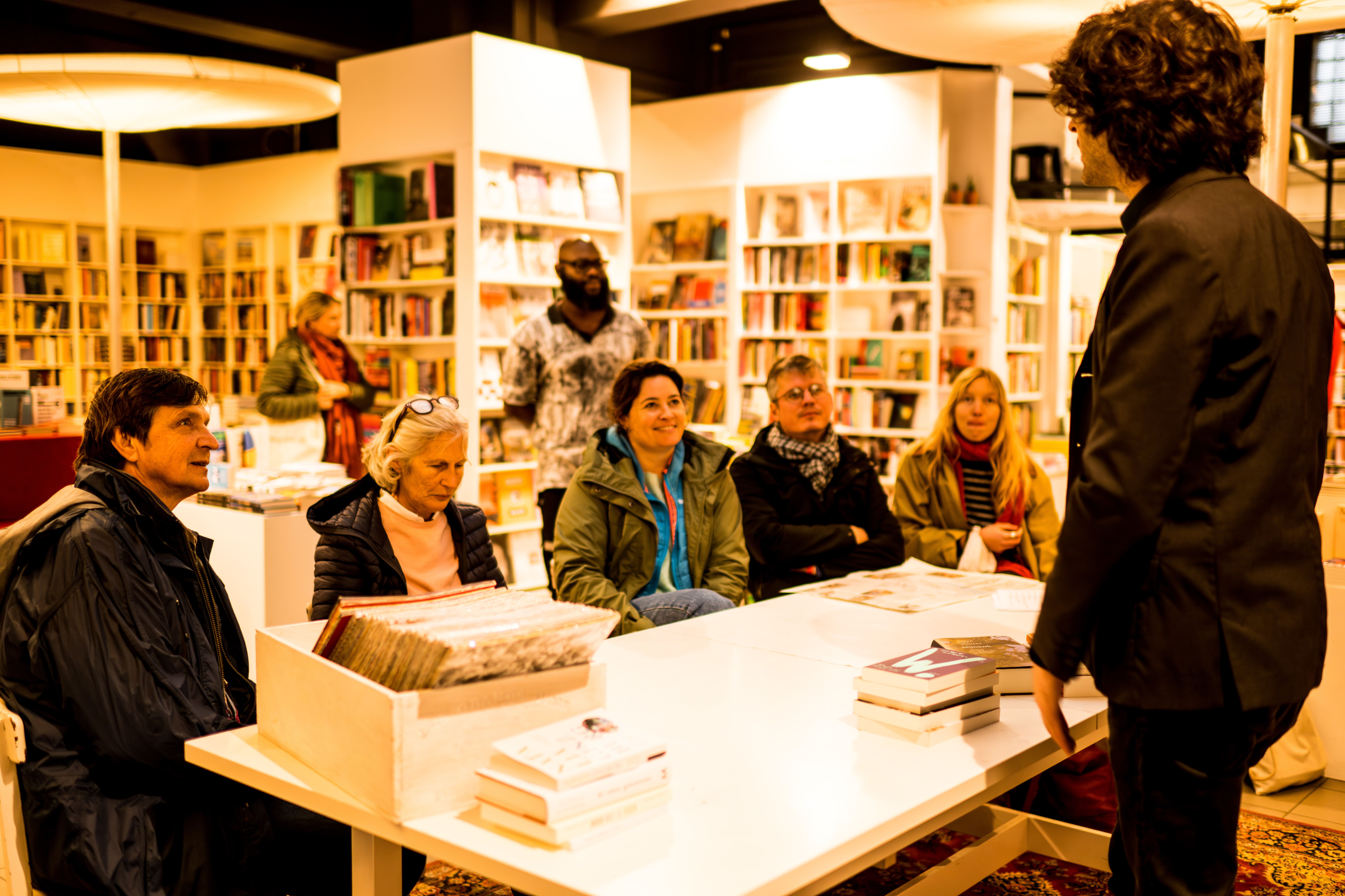 Een groepje mensen zit rond een tafel in een boekhandel. Aan de tafel staat ook een man recht die wat boeken vasthoudt en iets vertelt aan de groep.