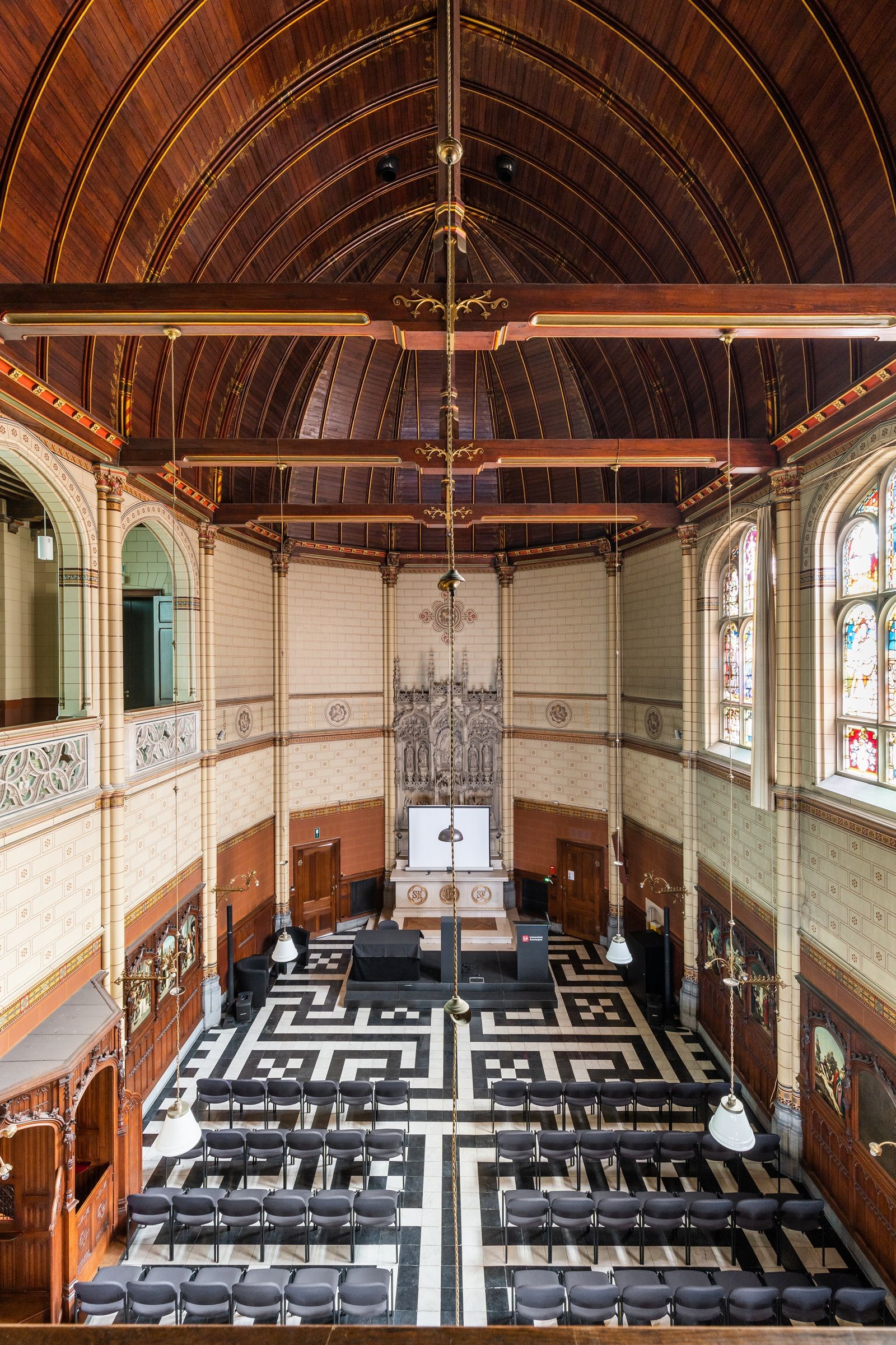 Houten plafond, witte muren met baksteenimitatie eronder een houten lambrisering en een vloer in geometrisch patroon met zwarte en witte tegels