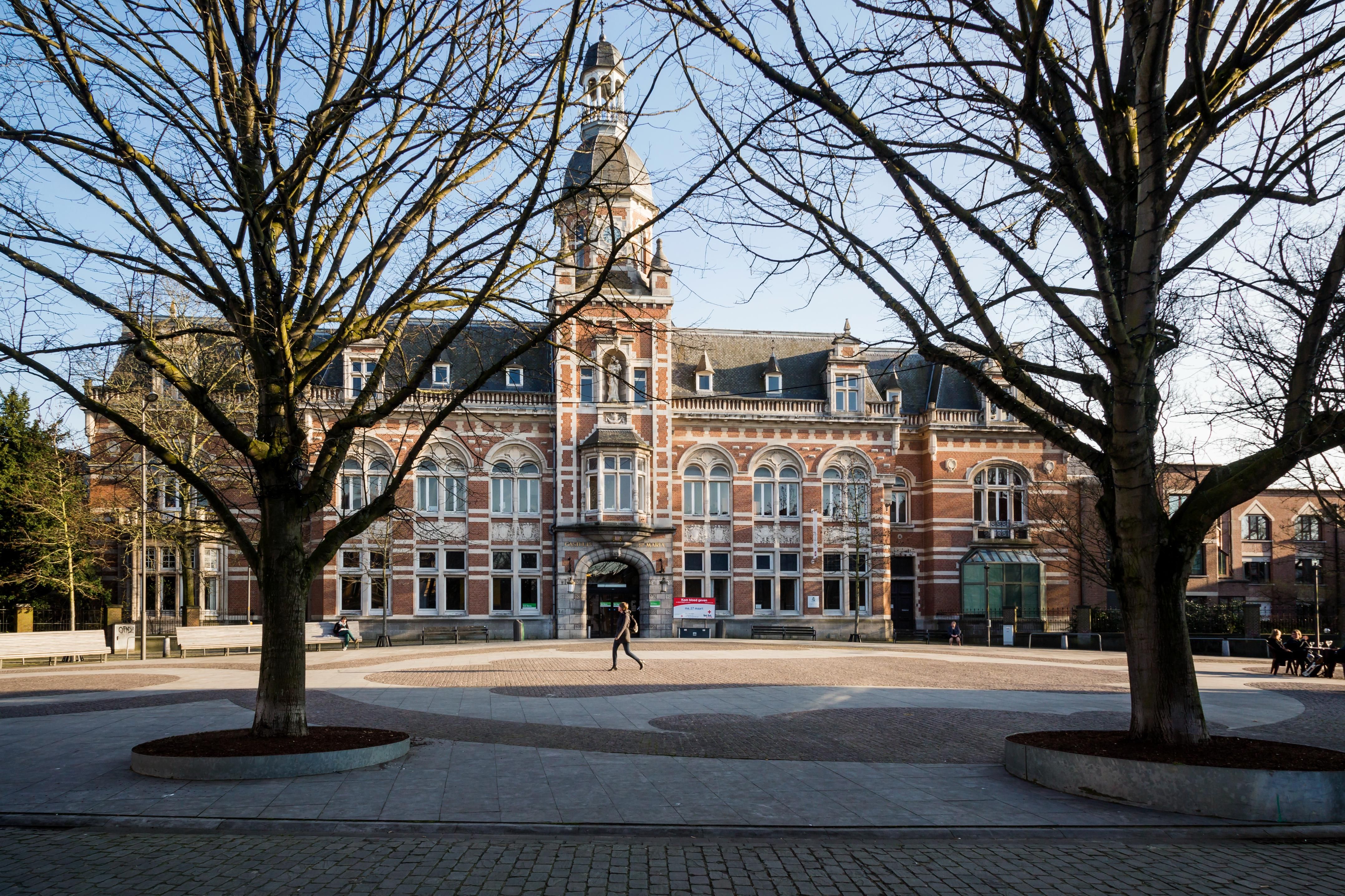 Het gebouw met toren volledig in beeld, vanop het plein met bomen ervoor.