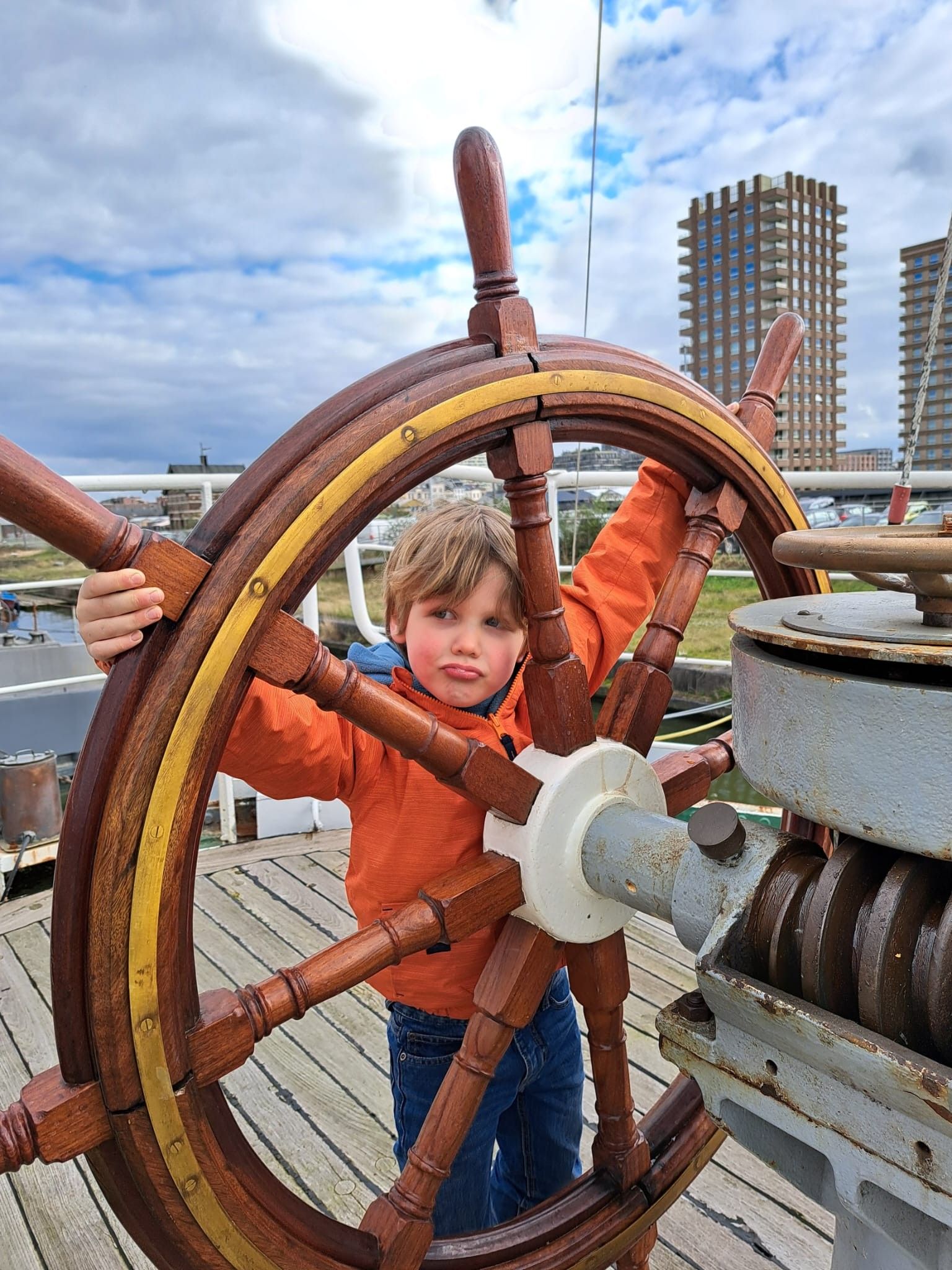 Jongen van ongeveer 5 jaar met oranje jas draait aan het roer van een schip
